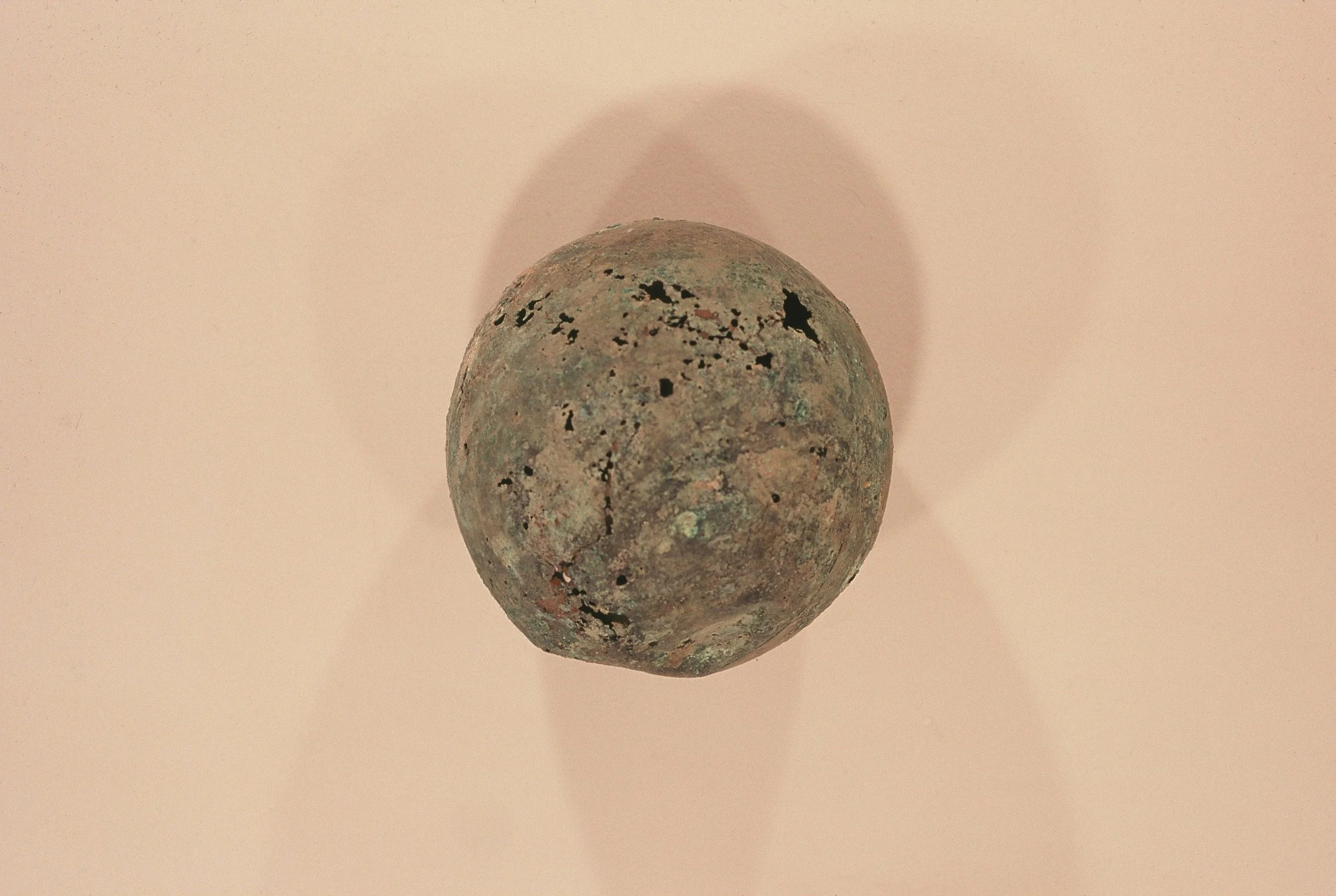 A close-up view of a small, round, greenish-black, weathered object with black spots, possibly a mineral or artifact, casting a shadow on a plain, light pink background.