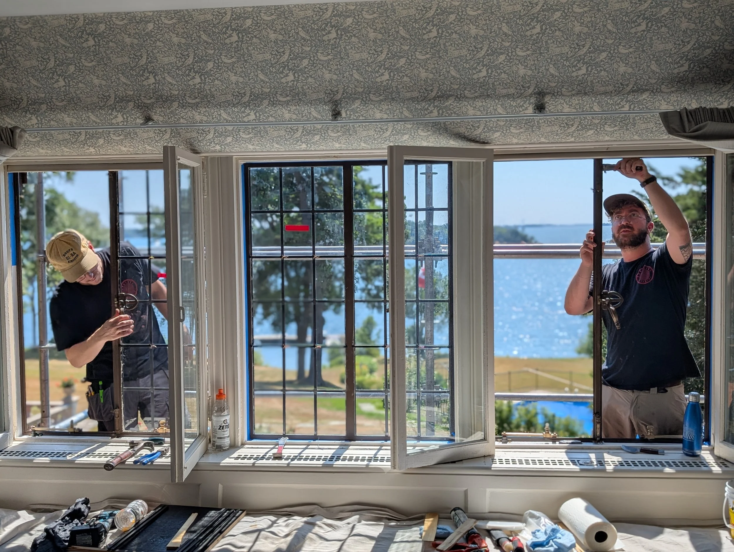 Two workers installing or repairing a window in a room with a view of a lake and trees outside. One worker is on the left outside the window, and the other is inside the room on the right, both working on the window frame.