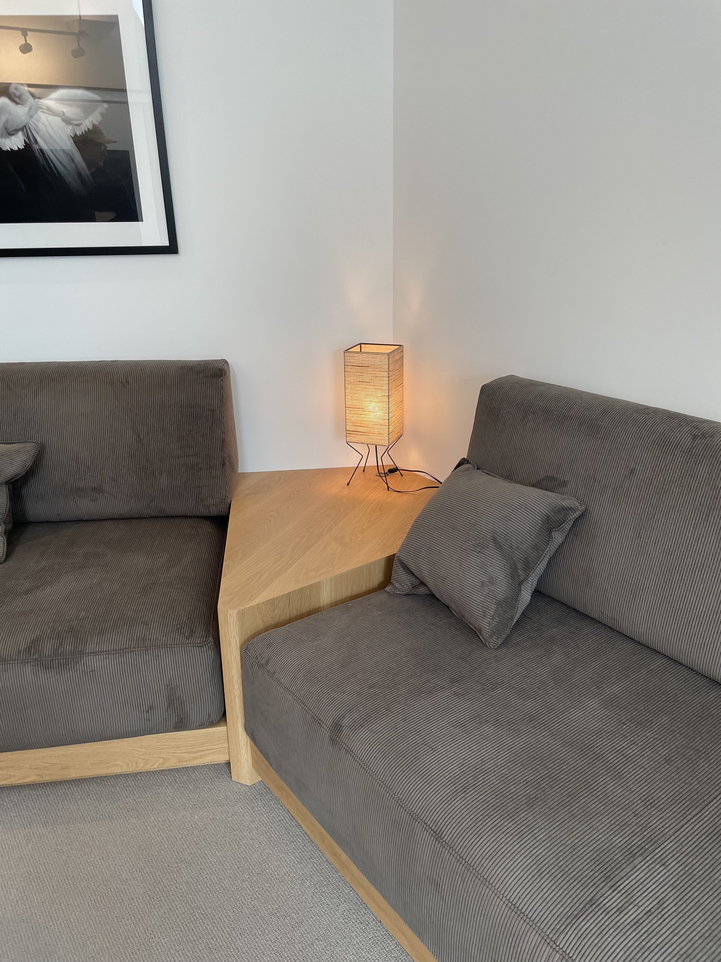 Living room corner with two gray upholstered couches separated by a wooden triangular side table, which holds a small illuminated table lamp. A framed black-and-white photo hangs on the white wall above the left couch.