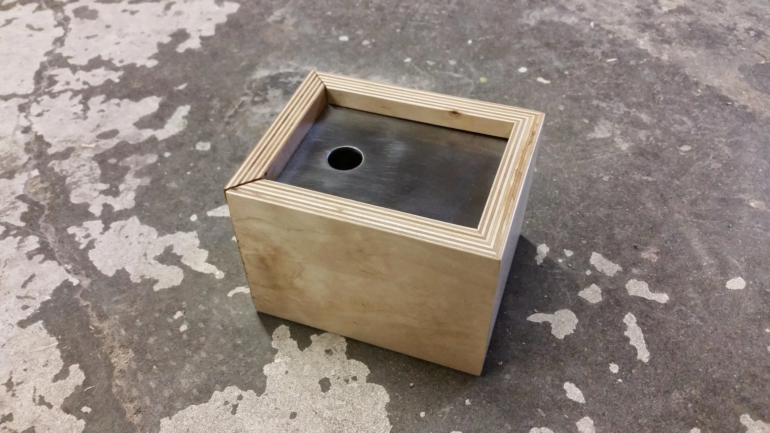 A small wooden box with a metal top and a round hole in the center, placed on a worn concrete surface.