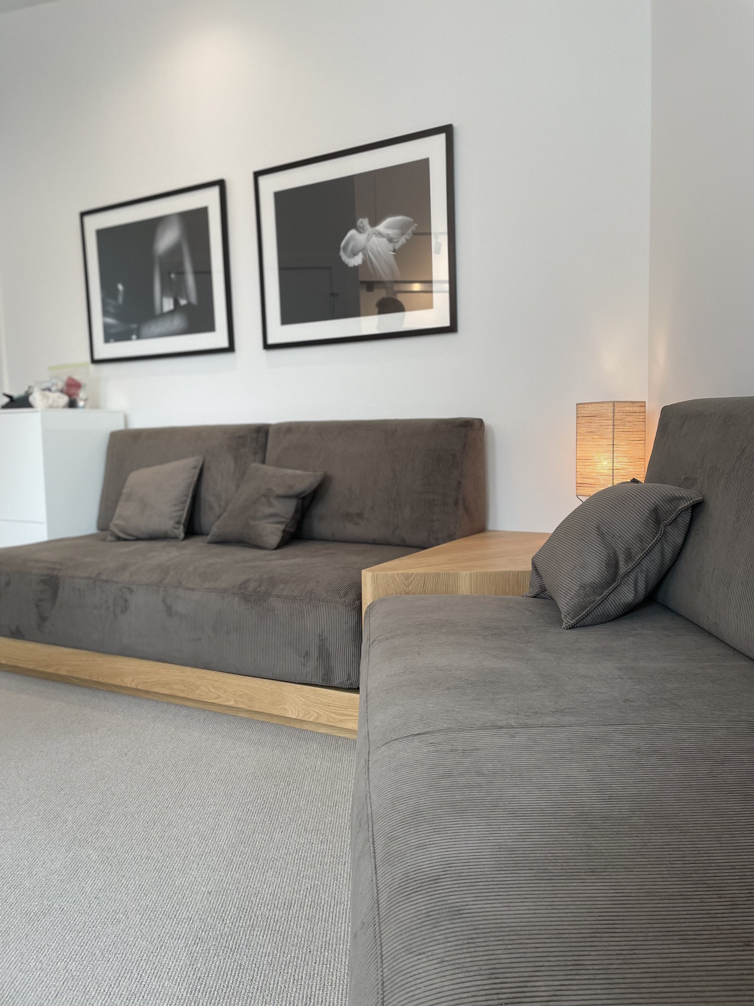Living room with gray sofas, black-and-white framed photographs on the wall, a wooden side table, and a bedside lamp.