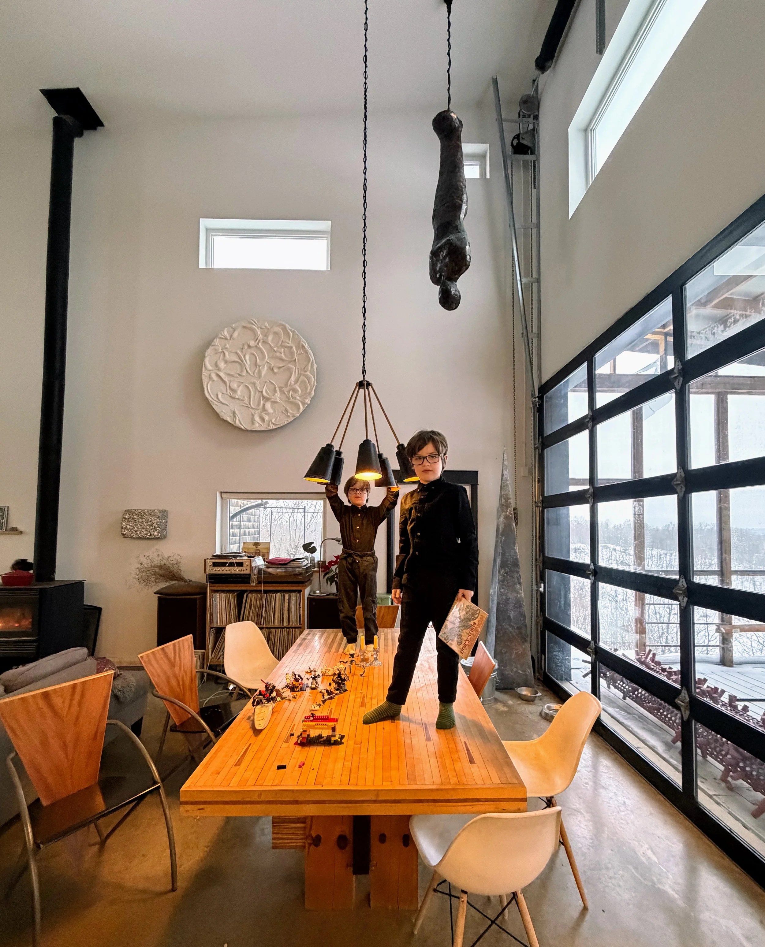 A modern dining room with two children standing on a wooden table, one holding a pendant light fixture, and toys scattered on the table. Large windows and an oversized garage door are visible, with snow outside.