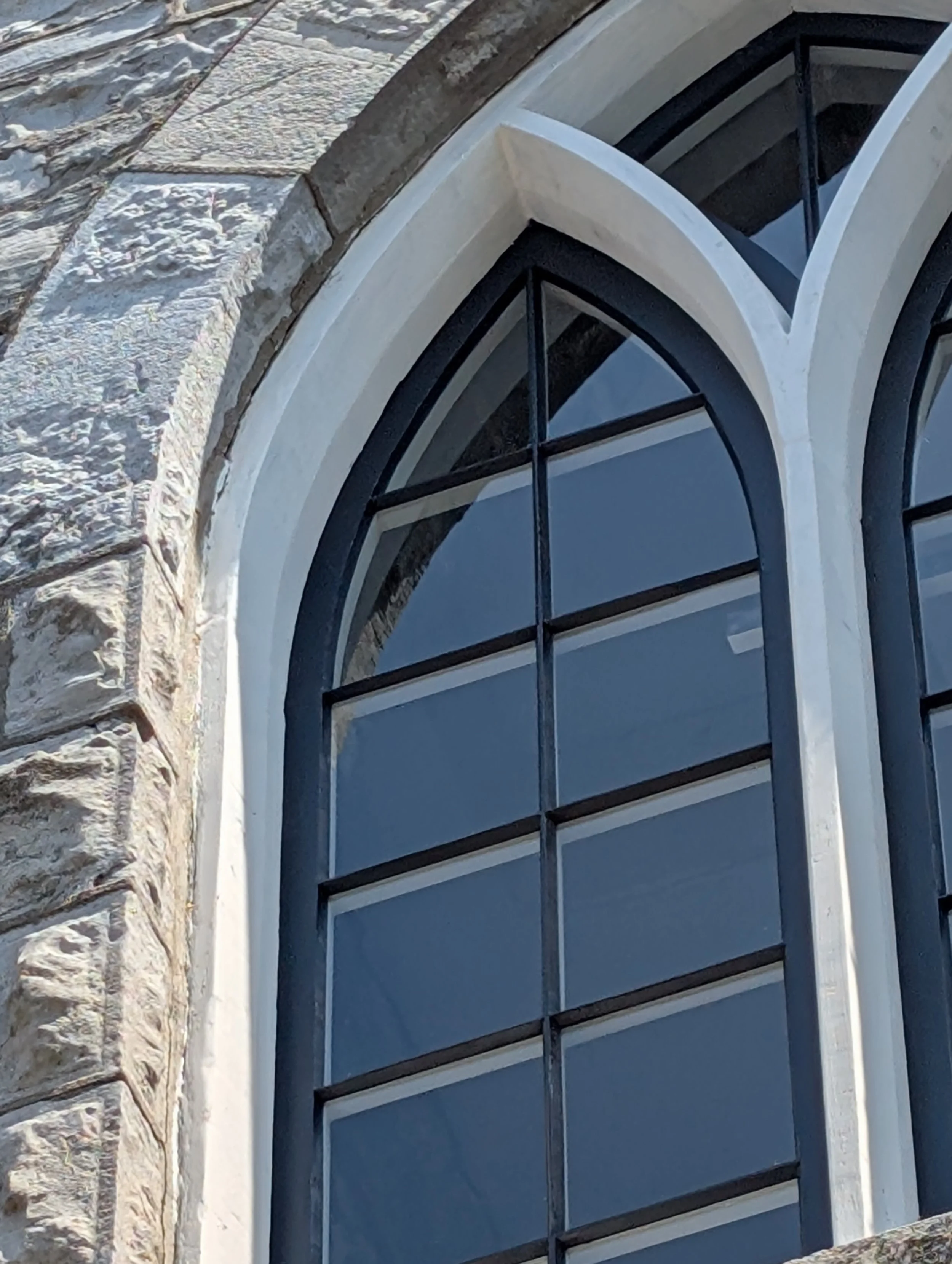 Close-up of a tall, arched Gothic-style window with black metal framing and white stone trim, set in a stone building wall.