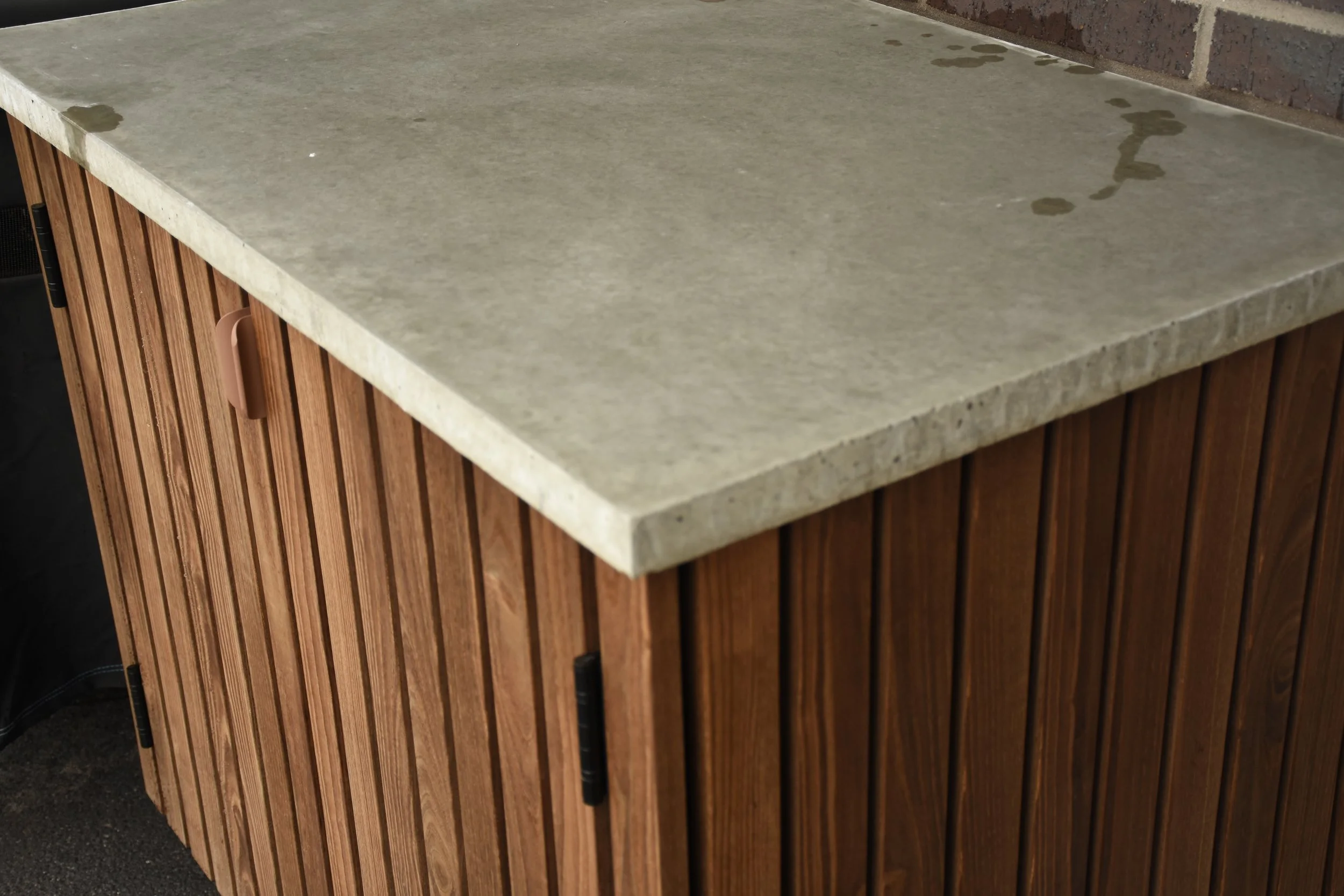 A wooden cabinet with vertical slats topped by a concrete countertop with some water spots.