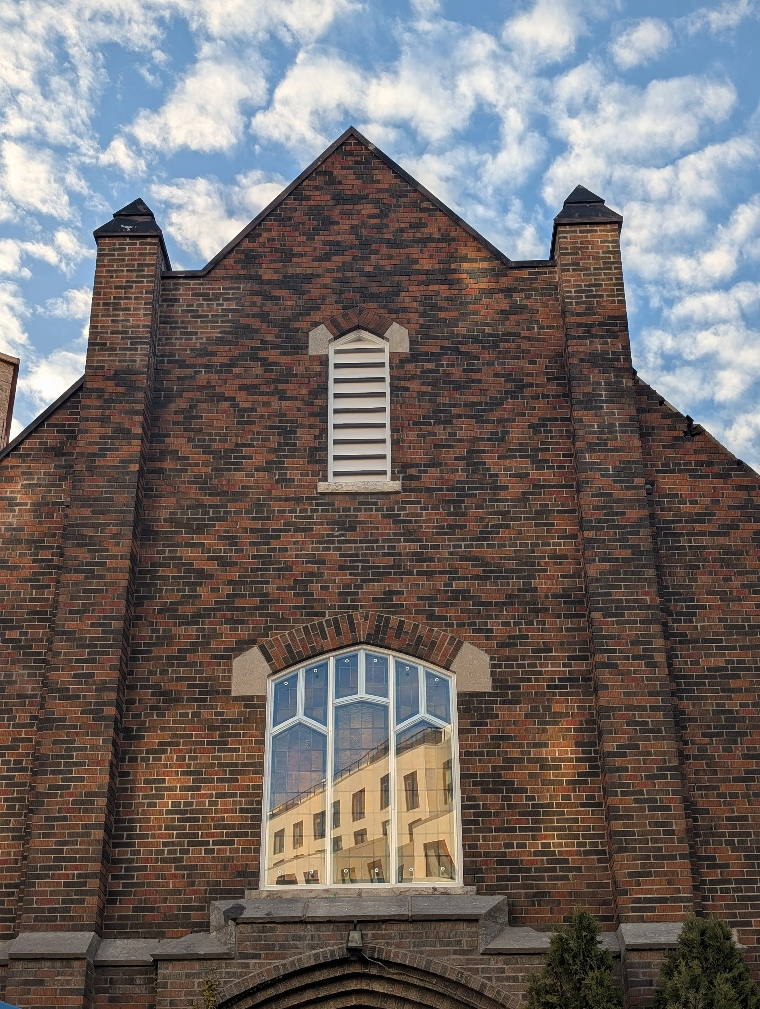 A brick building with a pointed roof and a large arched window reflecting a neighboring building, against a partly cloudy sky.