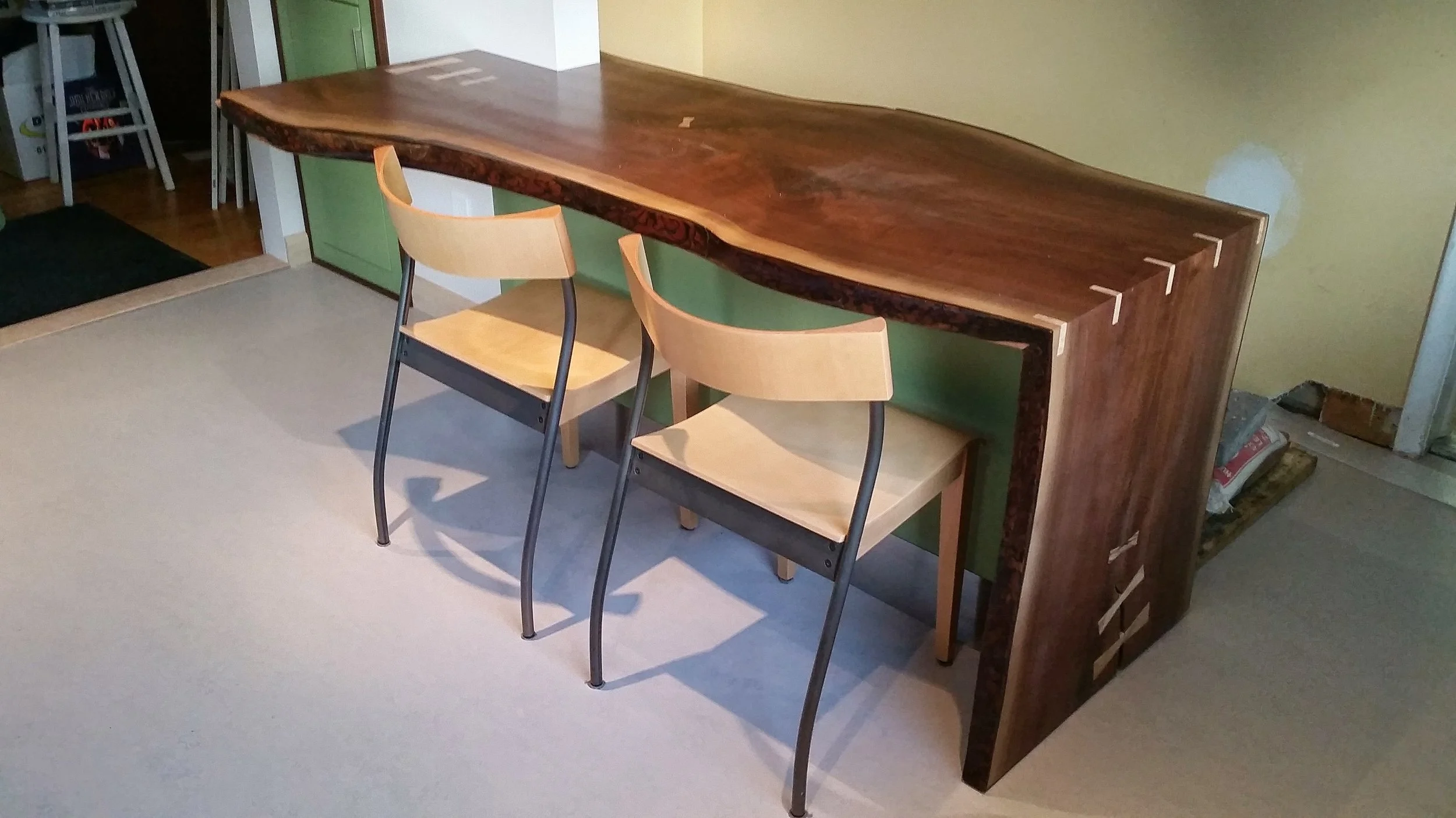 A rustic wooden bar-height table with natural edge, accompanied by two wooden chairs with black metal frames, in a room with a beige carpet and green wall cabinet.