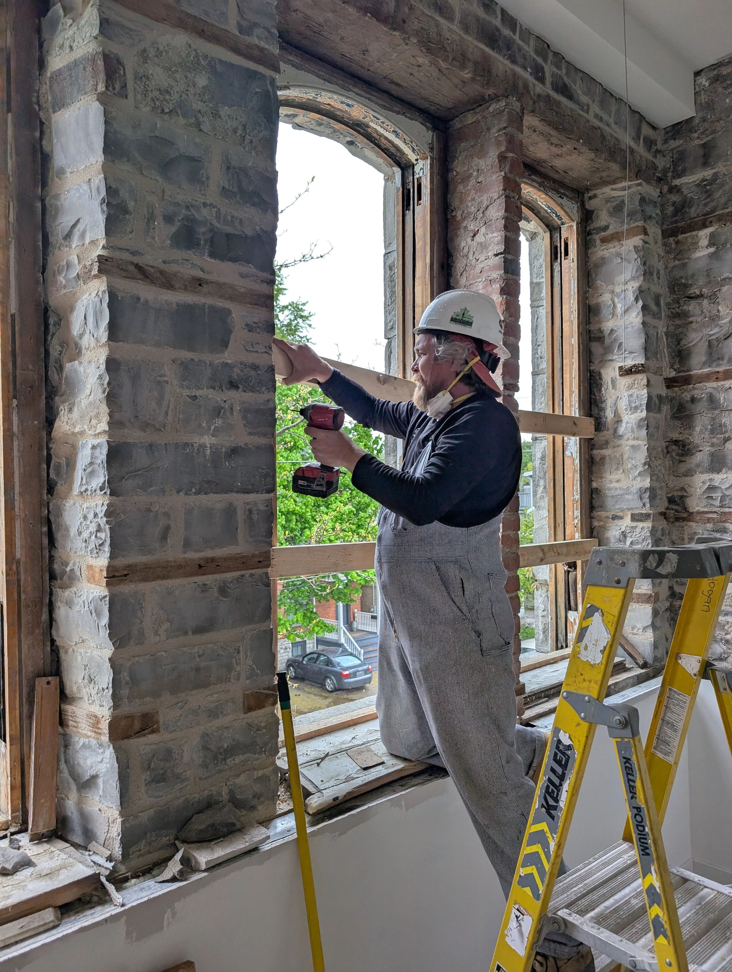 A construction worker wearing a white safety helmet and gray work clothes kneels on a step ladder while installing or repairing a brick wall next to a window in a building under renovation.