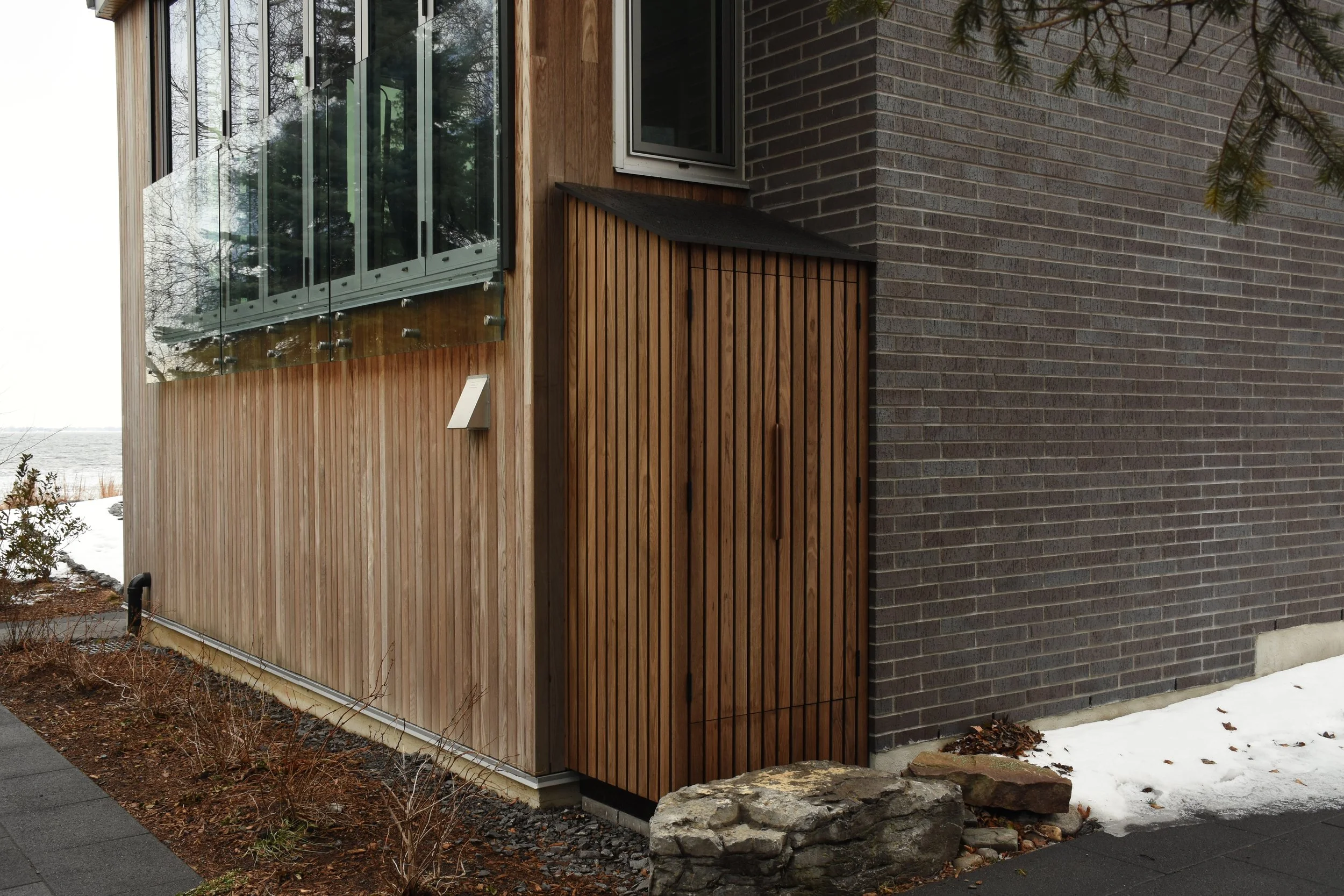 Close-up view of a modern house exterior with a mix of materials: wooden paneling, dark brick wall, and glass windows, in a snowy landscape.