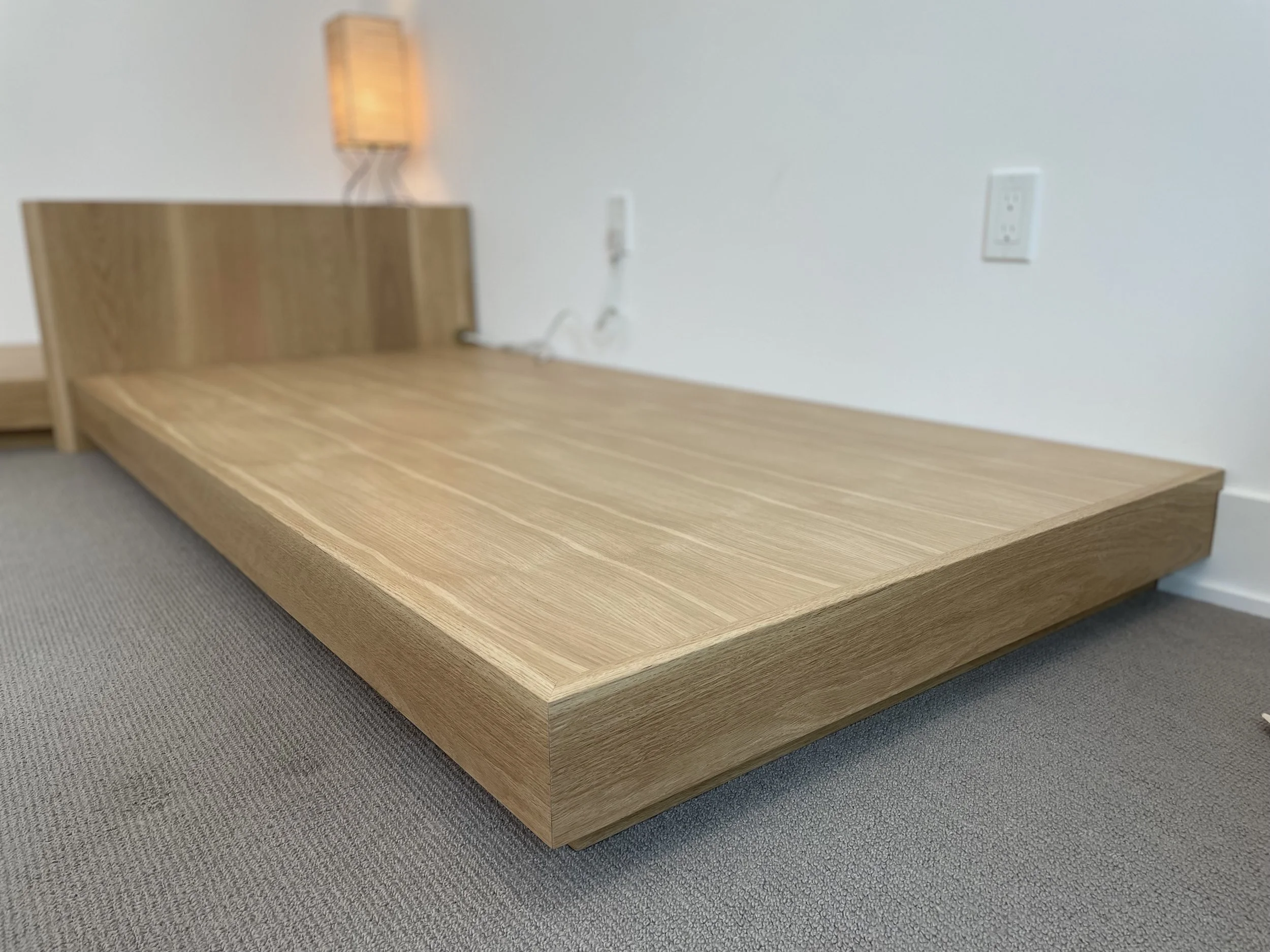 A low wooden bed frame with a headboard is placed on a beige carpeted floor. There is a wall with electrical outlets and a wall-mounted wooden lamp in the background.