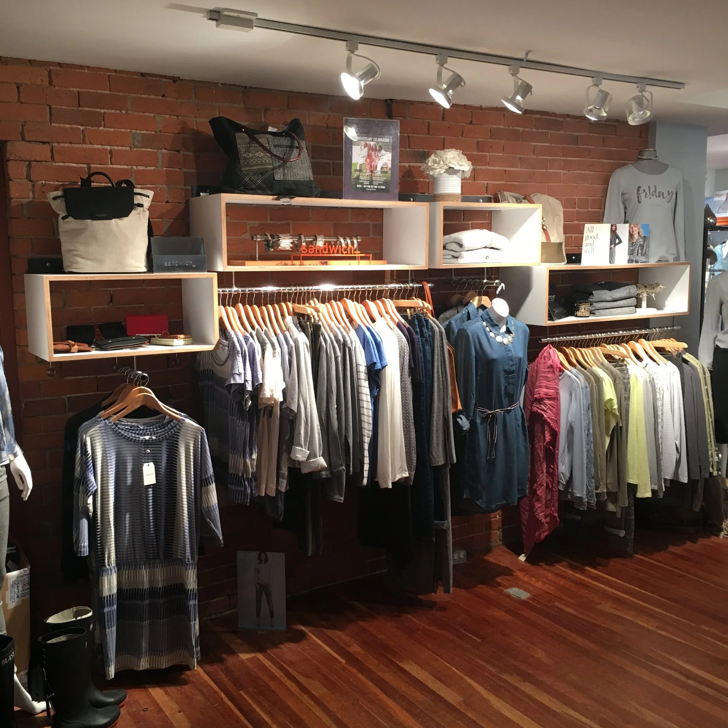 Clothing display in a retail store with racks of assorted women's apparel, handbags on shelves, and decorative signs against a brick wall.