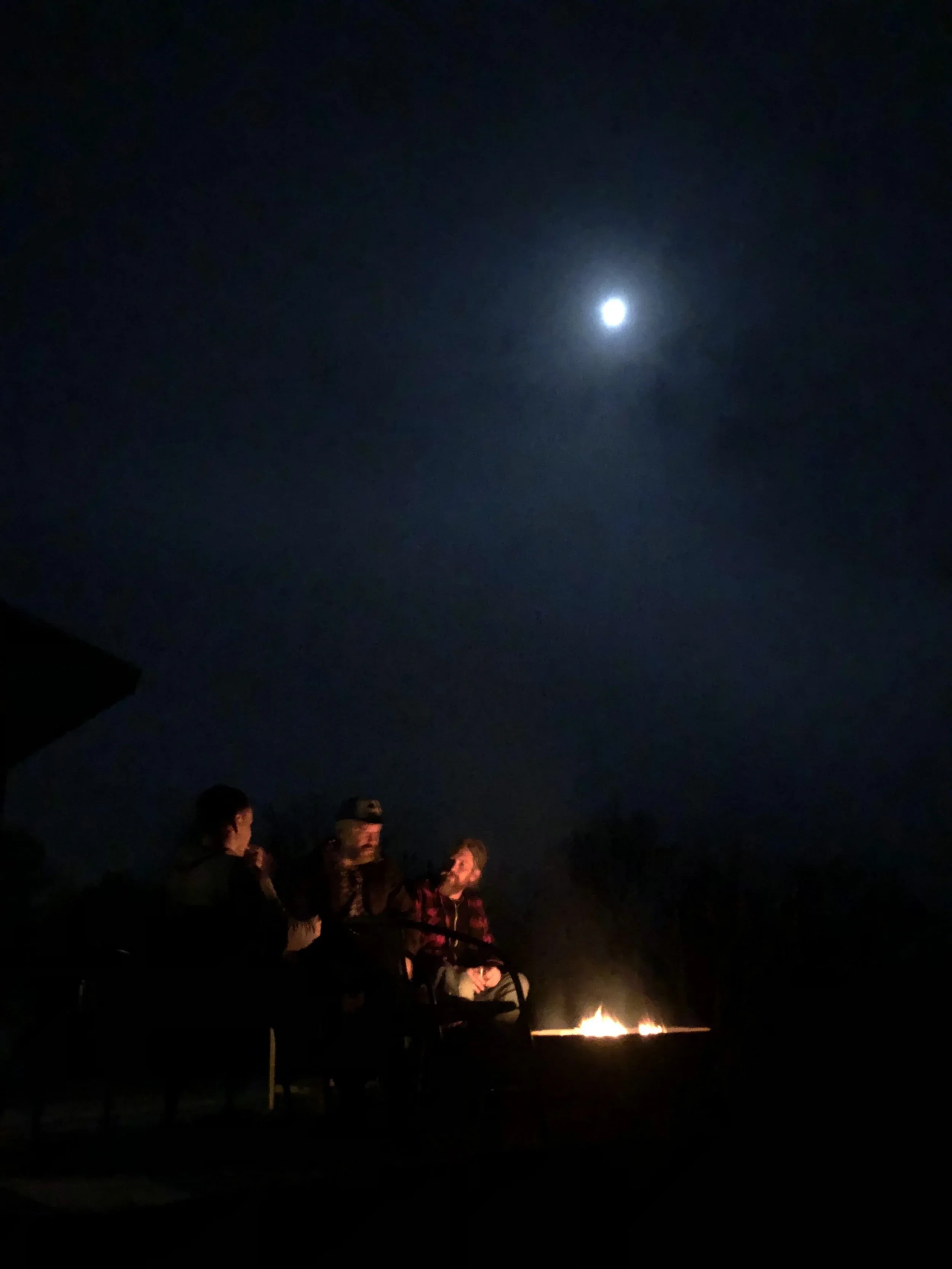 Three people sitting around a firepit outdoors at night with a bright full moon in the sky.