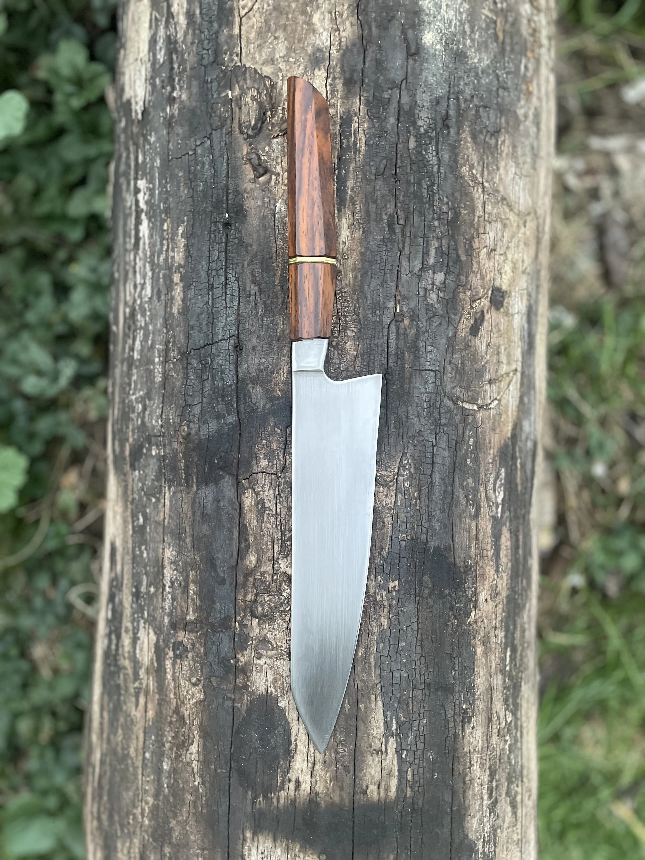 A large kitchen cleaver with a handle made of reddish wood and a brass band, resting on a weathered wooden surface outdoors.