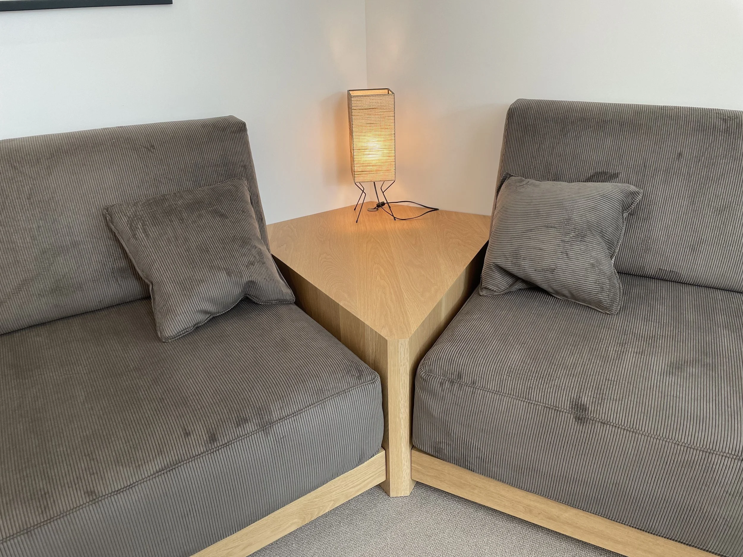 Two gray corduroy sofas with matching pillows, facing each other, separated by a light wood corner table with a small decorative lamp, in a neutral-colored room.