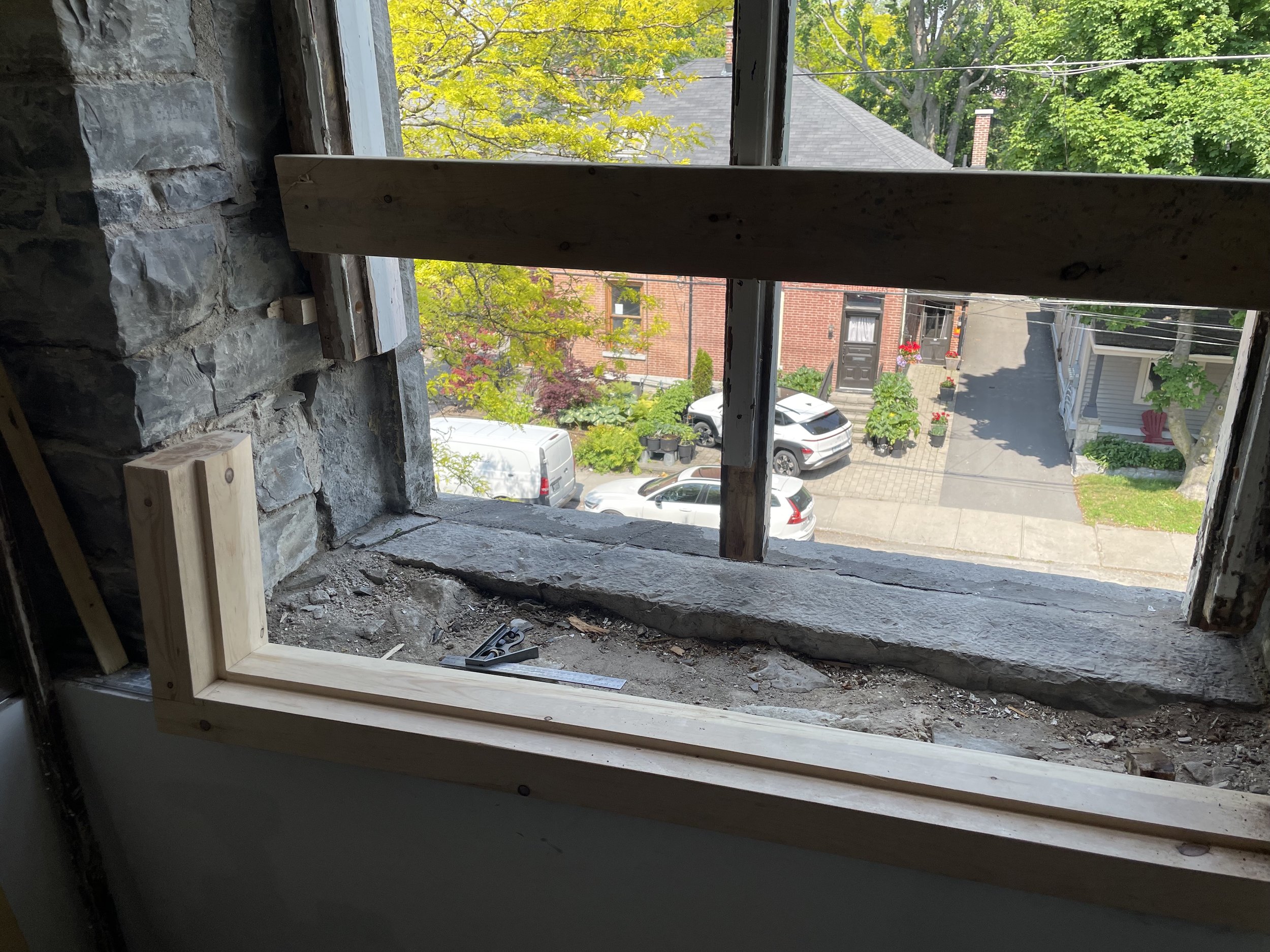 View through an unfinished window with wooden framing, overlooking a residential street with parked cars, brick houses, trees, and a sidewalk.