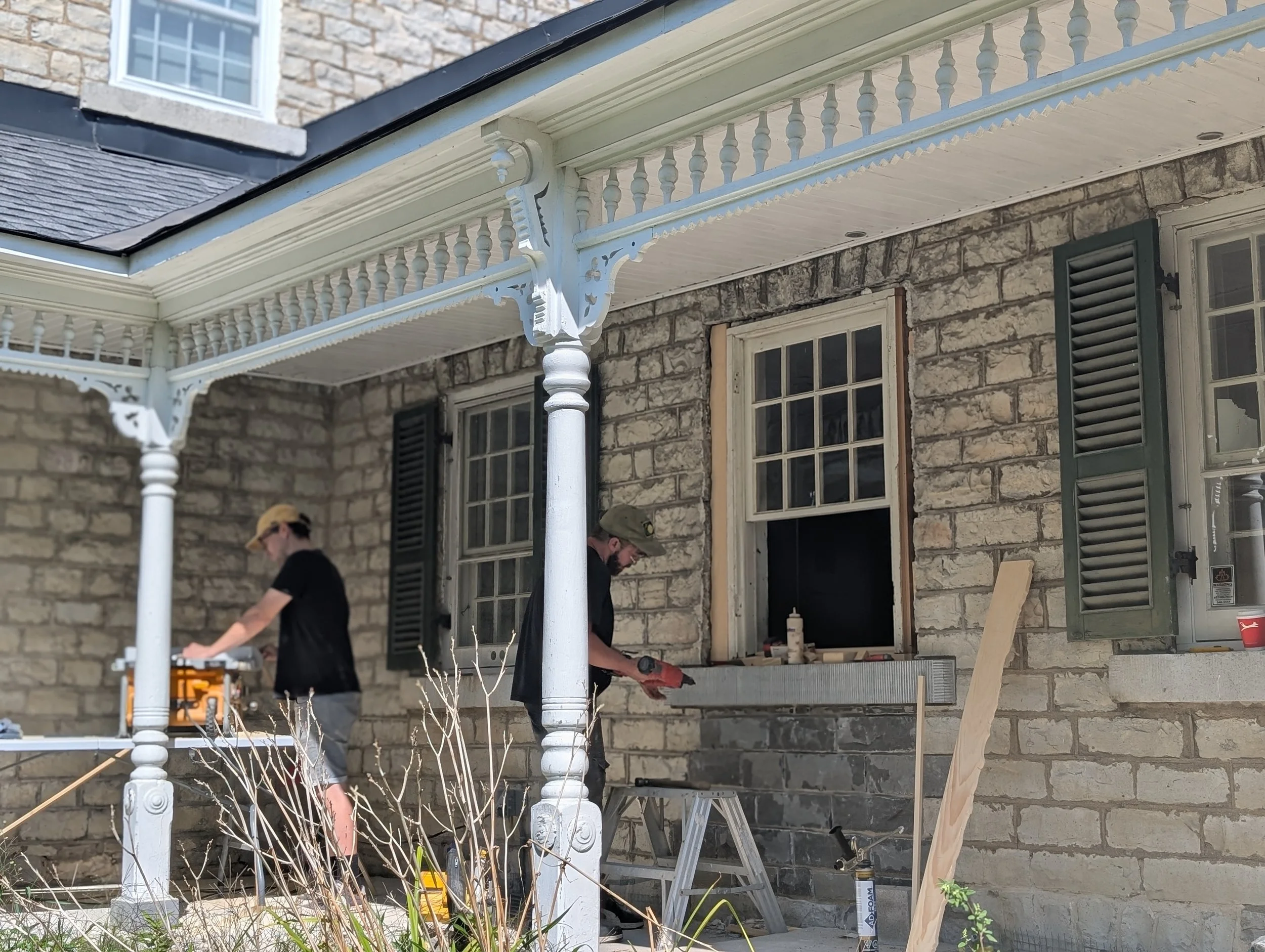 Two people working on a stone house front porch. One is using a saw, and the other is using a power tool. The porch has white decorative columns and trim, green shutters, and a window with an open black window pane. There are tools, a wooden board, a