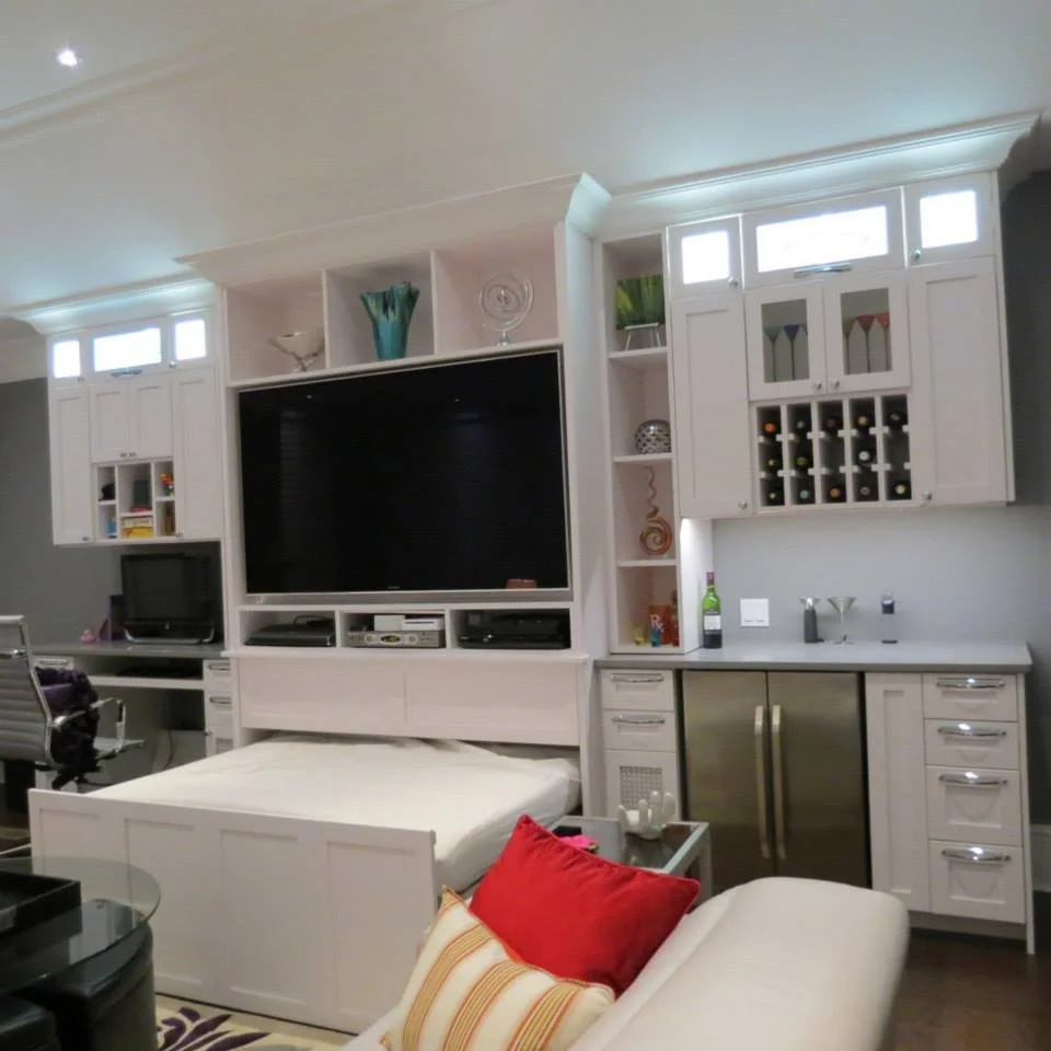 Living room with a large TV on a white built-in wall unit, a small desk with a computer, a bed with a white frame, and a seating area with a white sofa and red cushions.