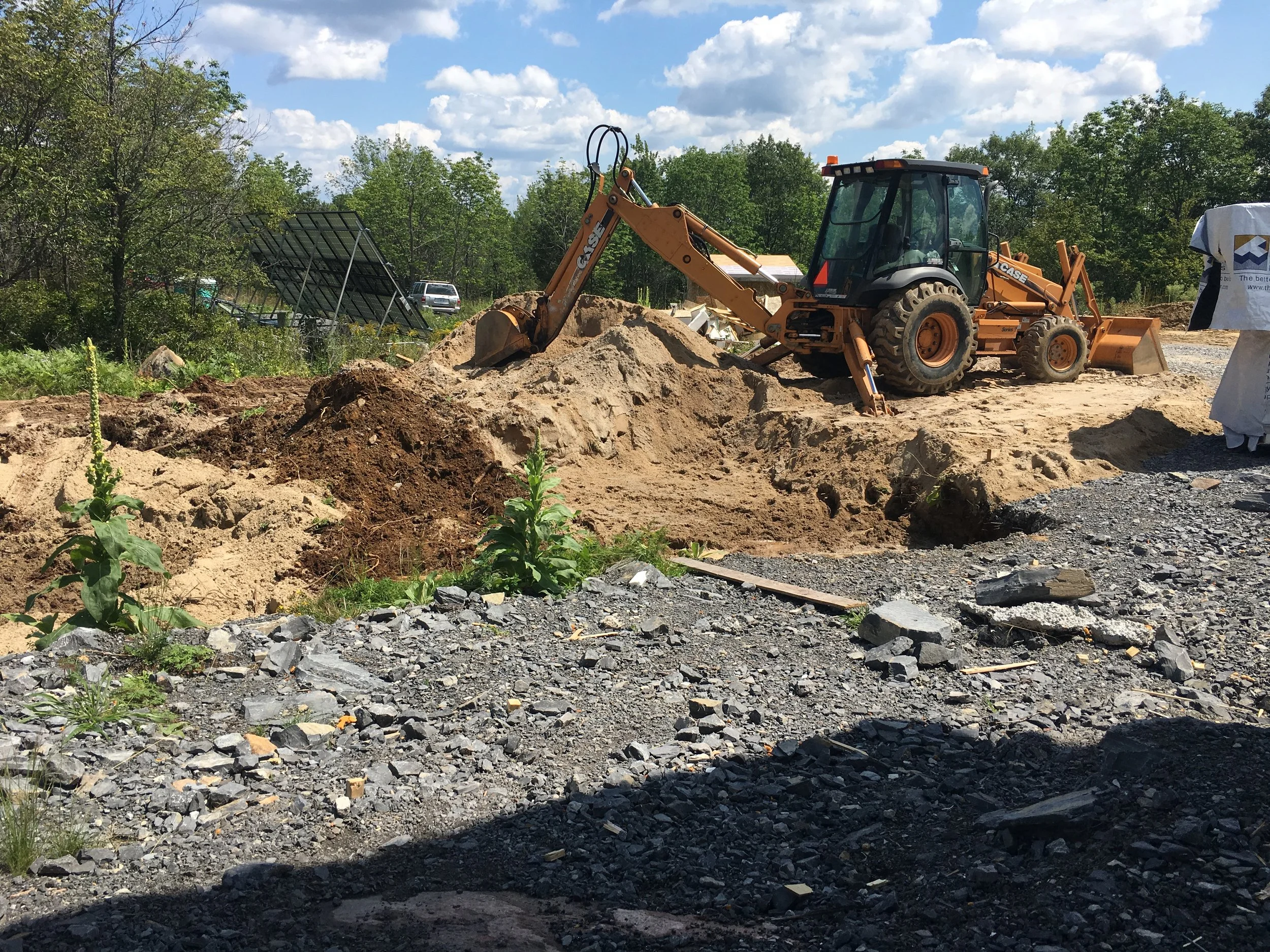 Construction site with a yellow backhoe digging into dirt, surrounded by gravel and some green plants, under a partly cloudy sky.