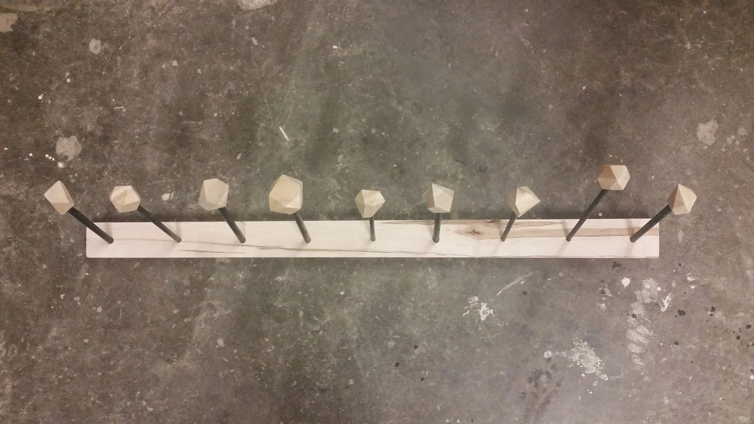 A wooden board with ten black nails, each topped with a beige, faceted, pentagon-shaped wooden knob, arranged in a straight line on a concrete floor.