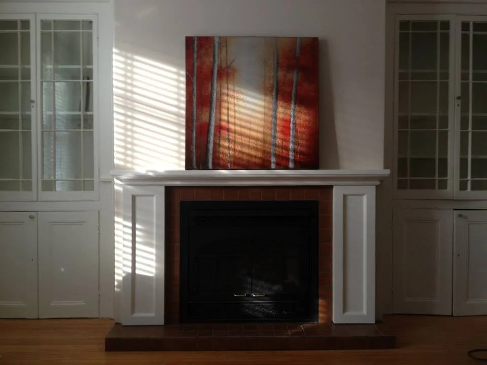 Living room with a white fireplace, abstract red and orange painting above it, and built-in white cabinets with glass doors on either side. Sunlight streams through window blinds, creating striped shadows.