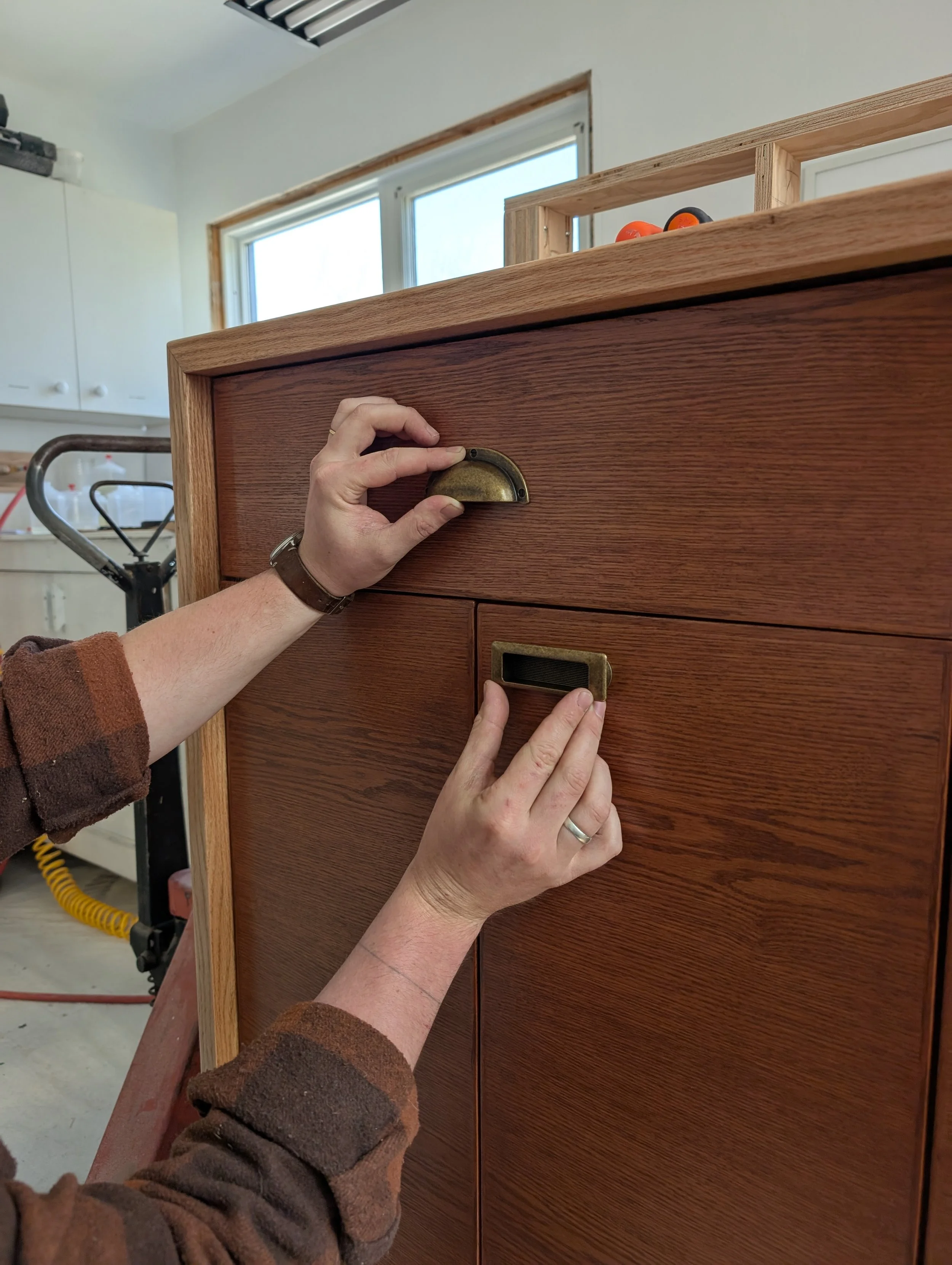 Person installing a vintage brass drawer handle and label holder on a wooden cabinet in a workshop.