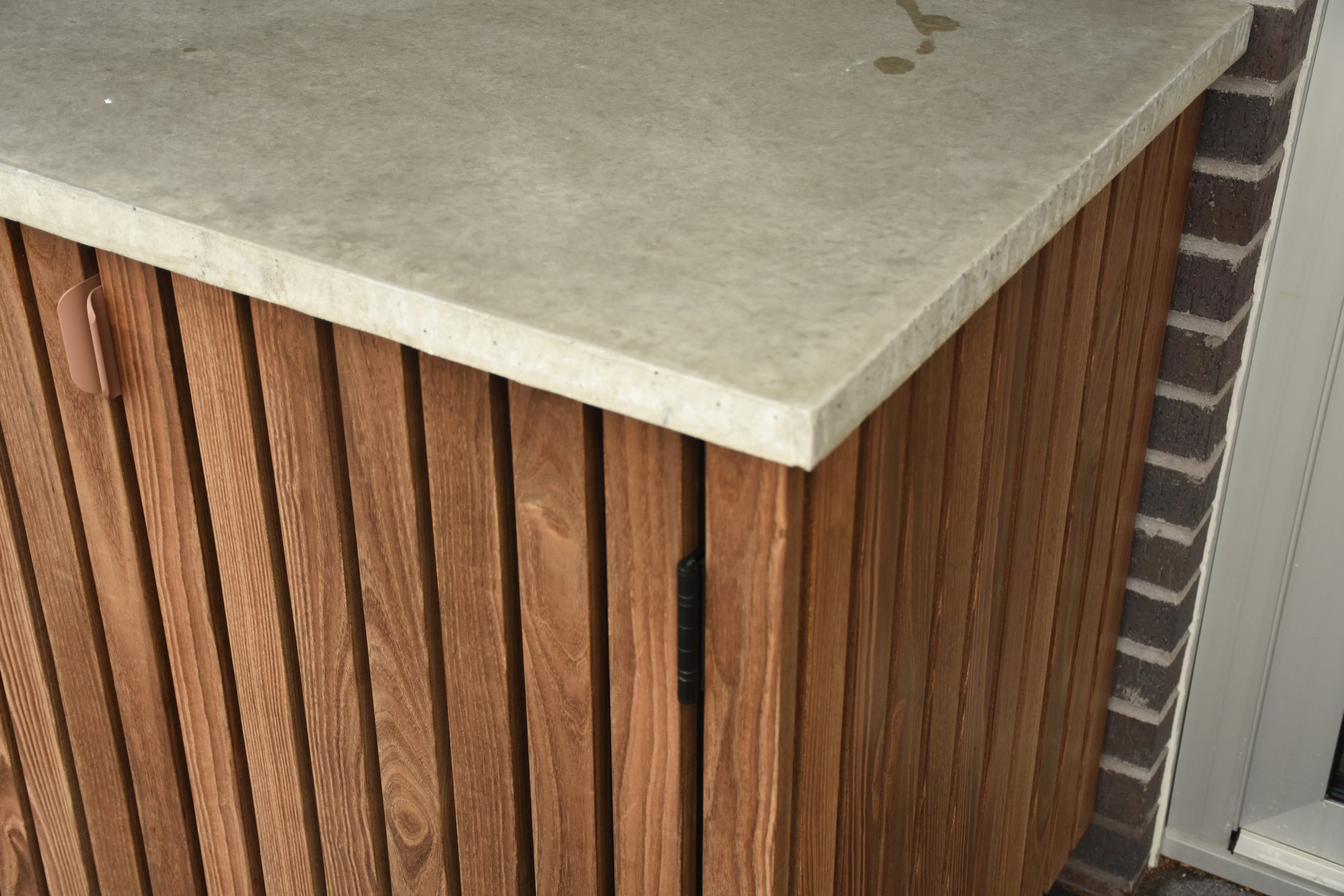 Close-up of a wooden cabinet with vertical slats and a beige-colored stone countertop next to a brick wall.