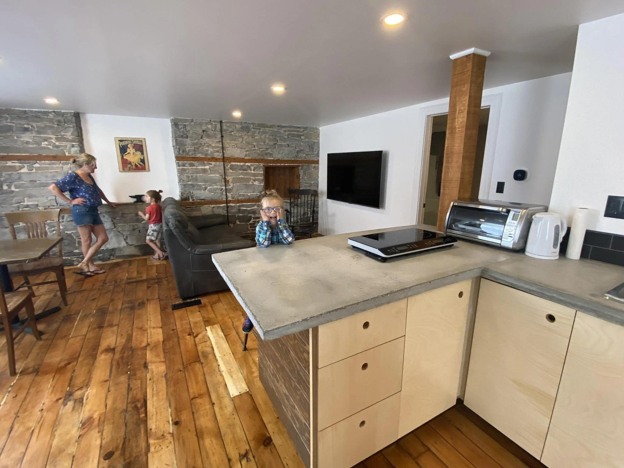 A family living room with a stone wall, wooden floors, and a kitchen counter in the foreground. A woman and two children are near the stone wall, with one child sitting on a black sofa and the other standing nearby. A woman stands next to the childre