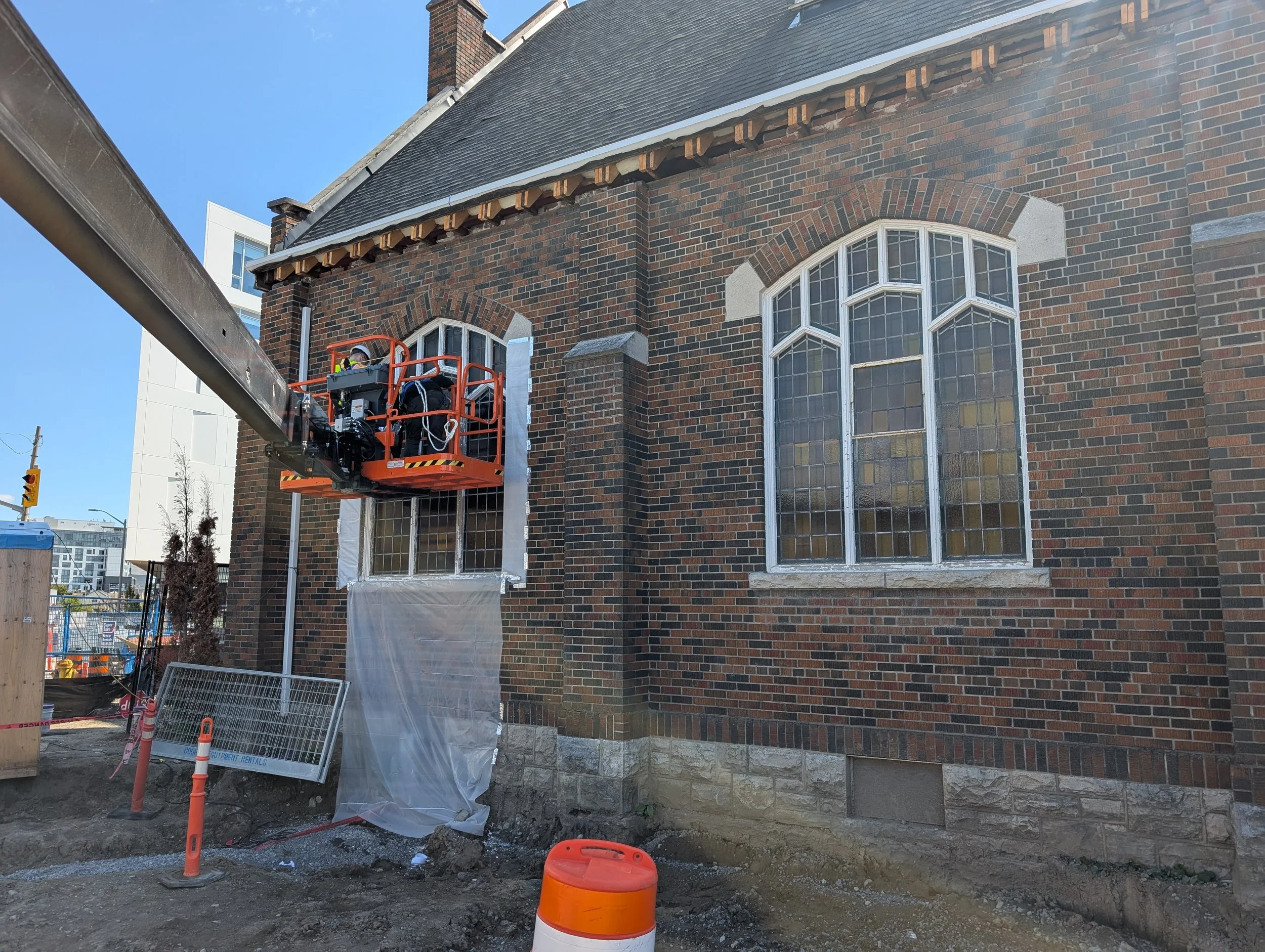 Construction worker in an orange lift working on a brick building's window, with construction barriers and equipment around.