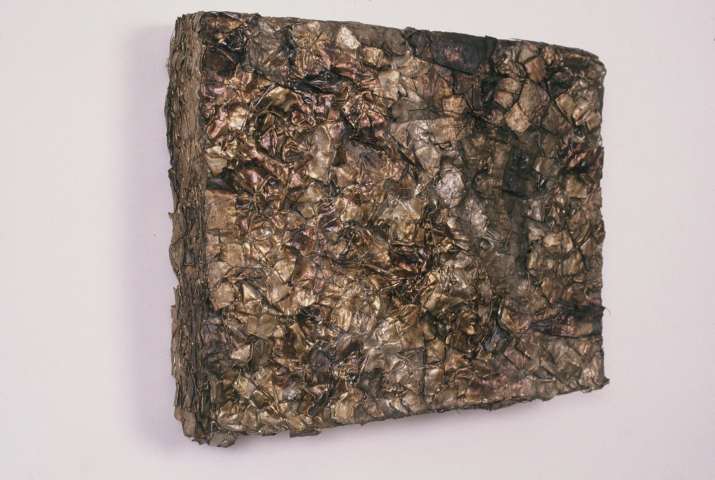 A close-up of a textured, metallic-looking, crumpled square artifact with irregular surface details and dark coloration.
