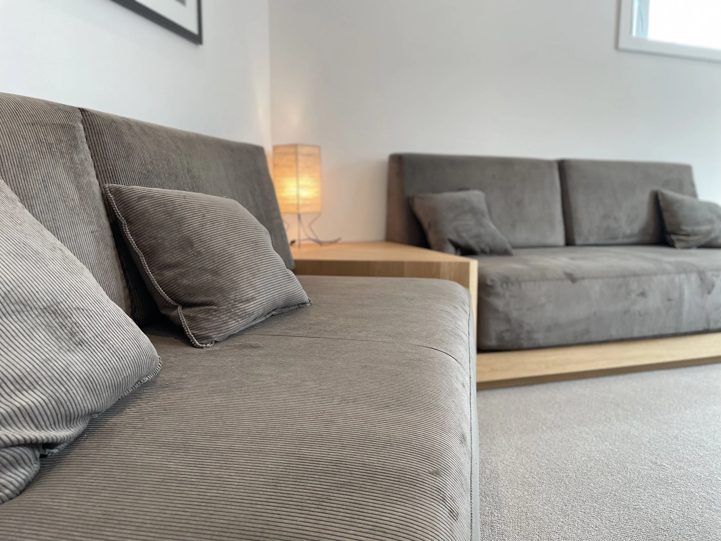 A living room with two grey sofas and a wooden side table between them. A small lamp sits on the side table, turned on, casting a warm glow. The room has a neutral-colored carpet and white walls.