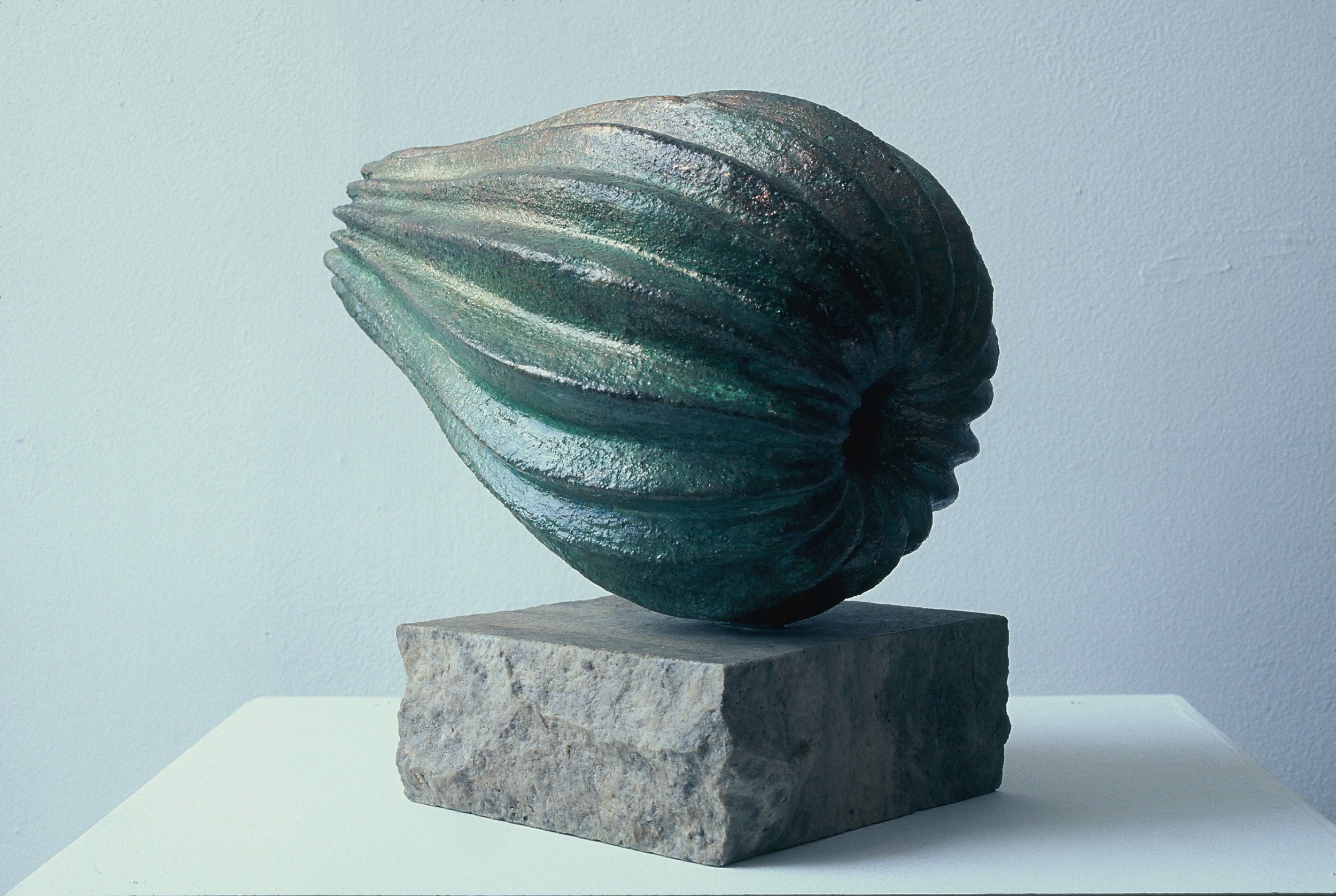 A modern sculpture of a stylized shell with ridges, mounted on a rectangular stone base, displayed against a plain light background.