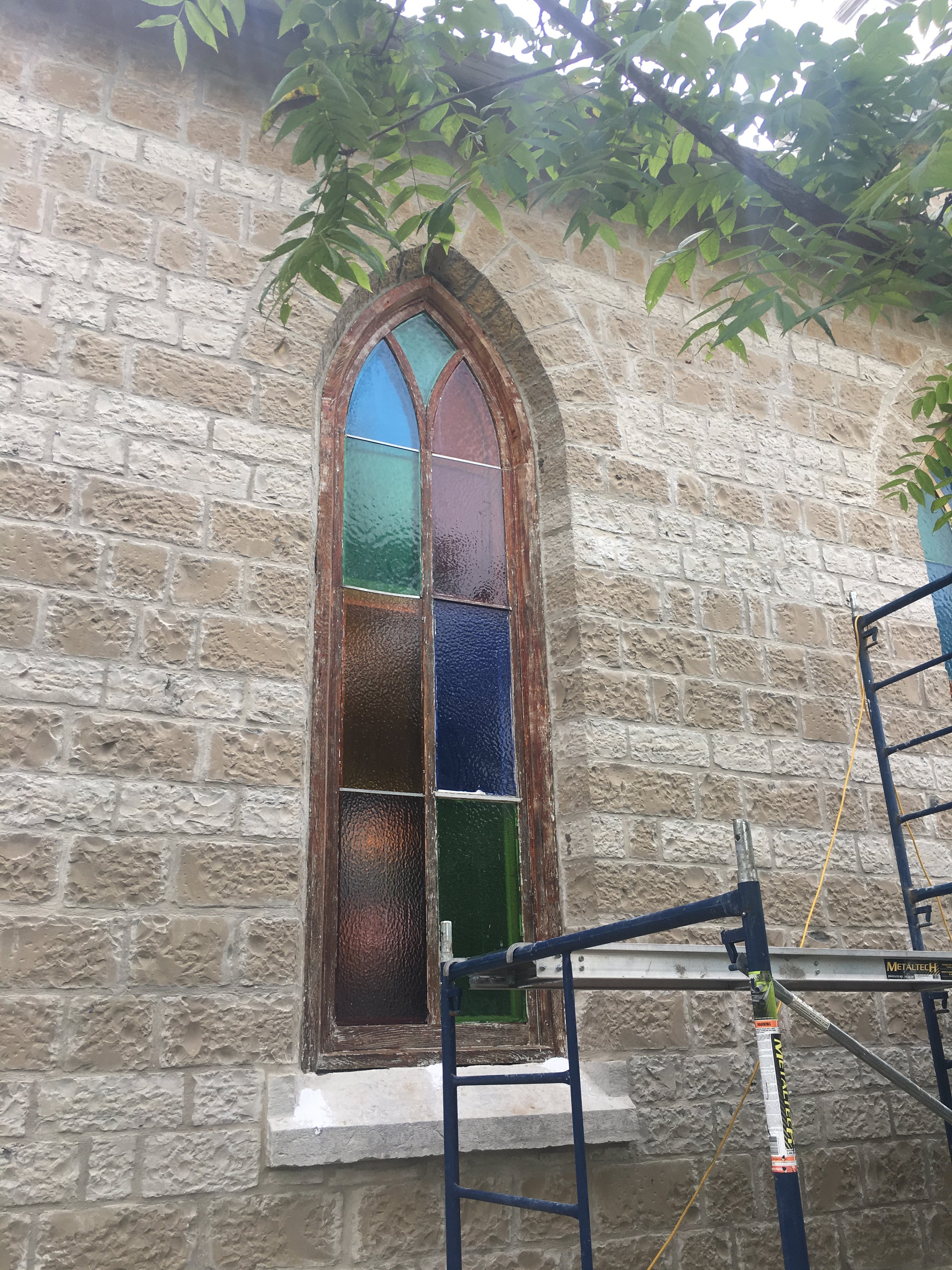 Colorful stained glass window on a stone church wall with scaffolding in front.
