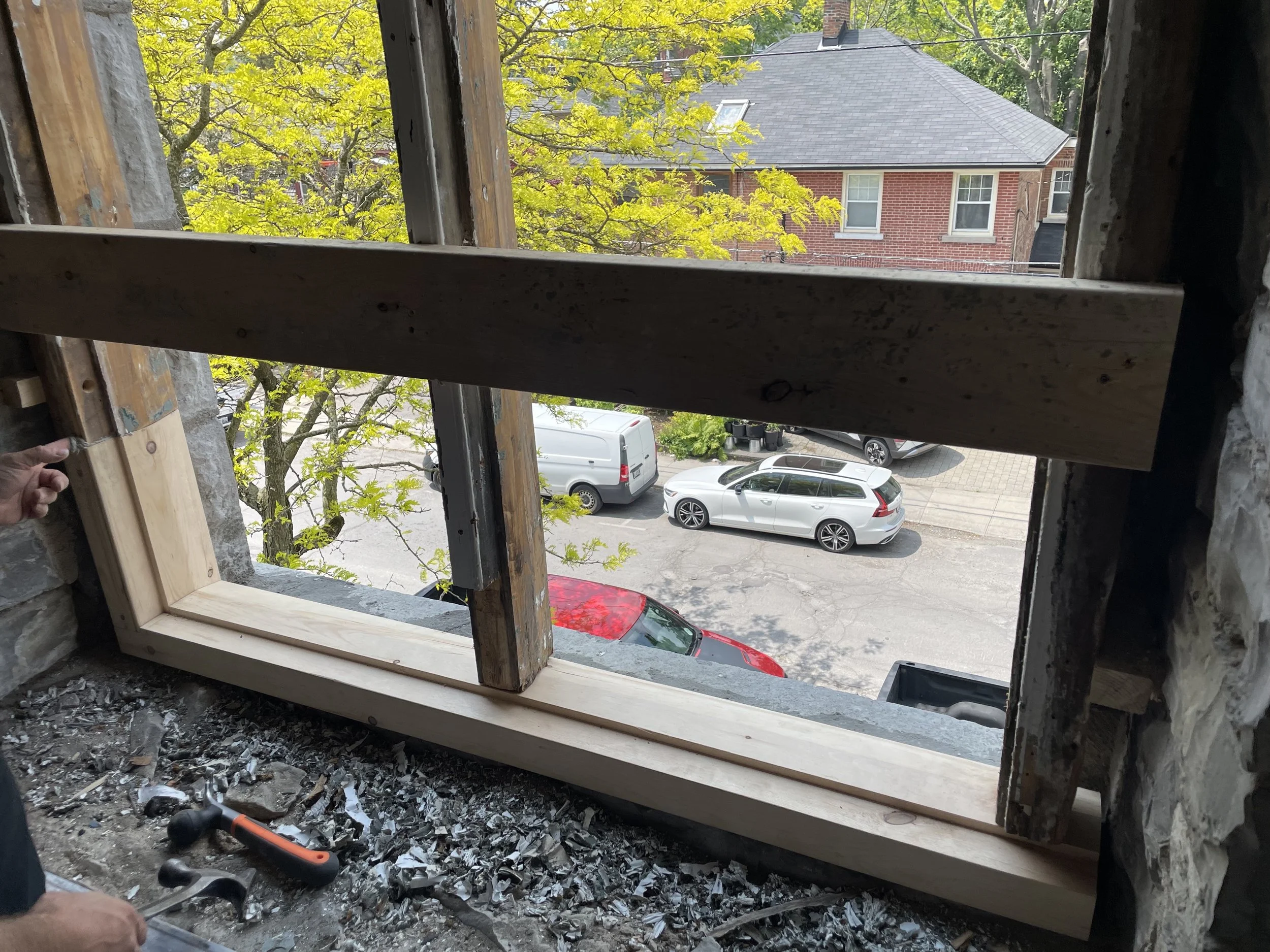 View from inside a building under construction, looking through a wooden-framed window opening, showing a street with parked cars and a red brick house across the street.