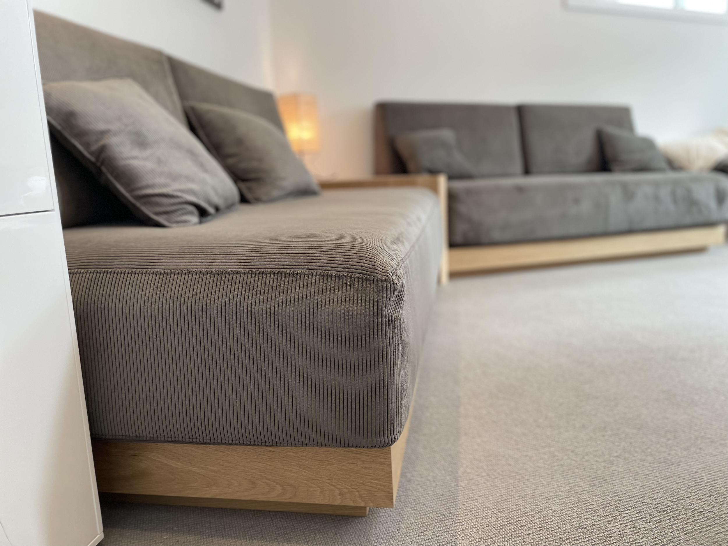 Close-up of a gray fabric sofa with gray pillows, in a living room with a beige carpet, a wooden side table, and a lit lamp in the background.