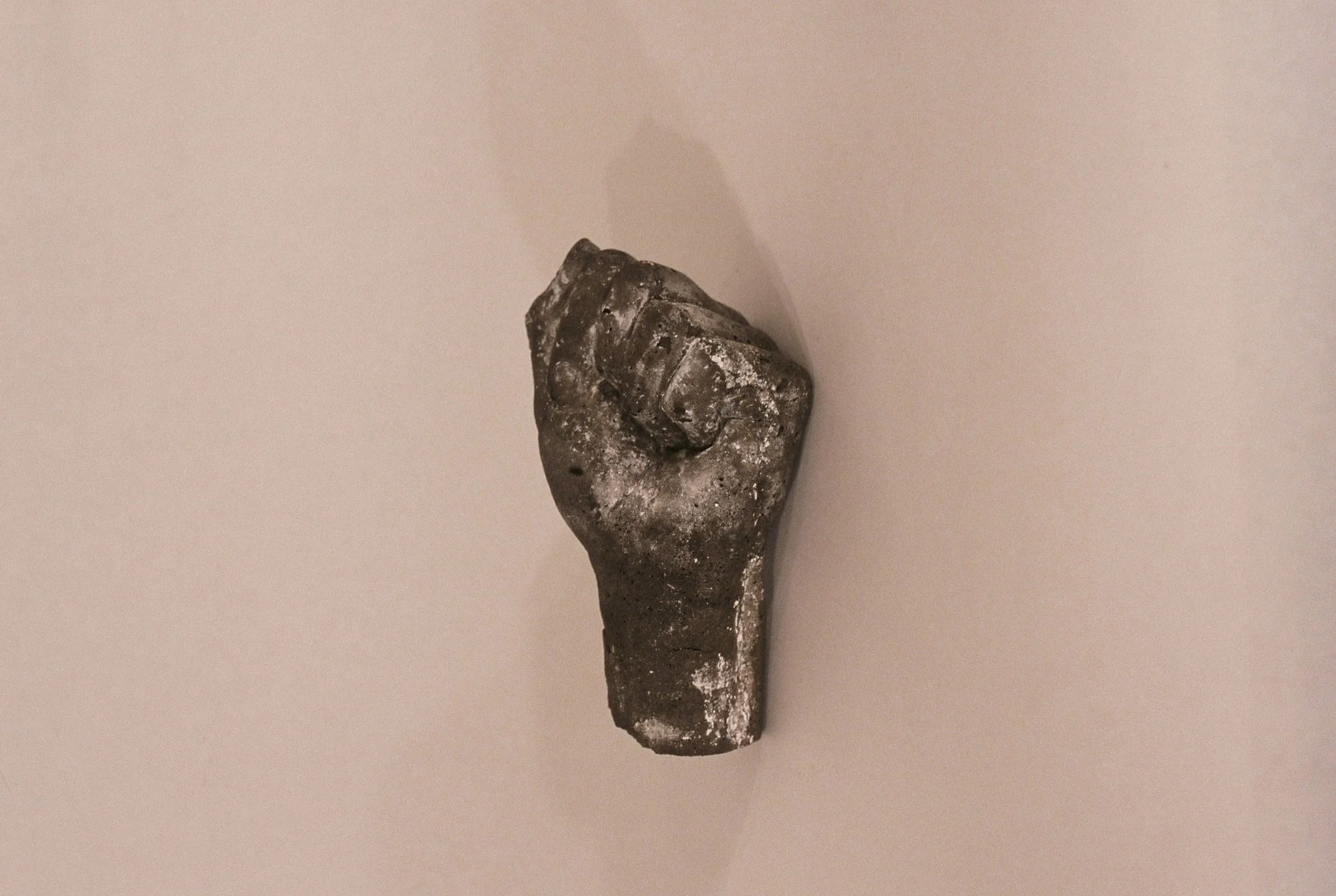 A small, dark-colored statue of a clenched fist with detailed fingers, mounted on a beige wall, casting a shadow to the left.
