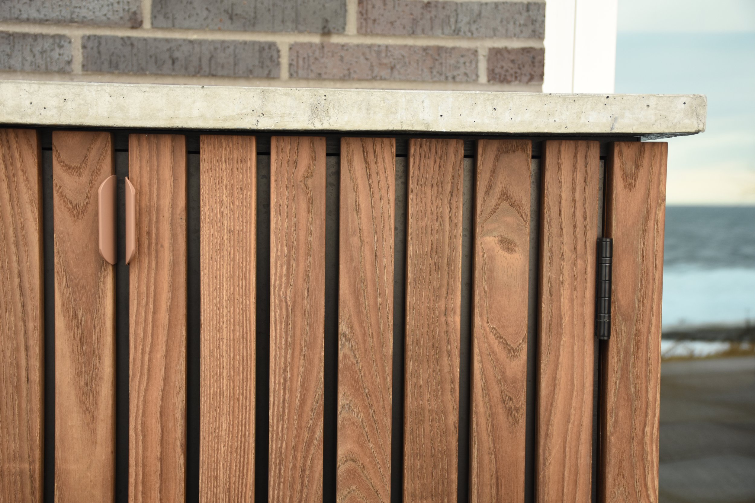 Close-up of a wooden cabinet with vertical slats, a clamp on the left side, and a hinge on the right, with an outdoor ocean view in the background.