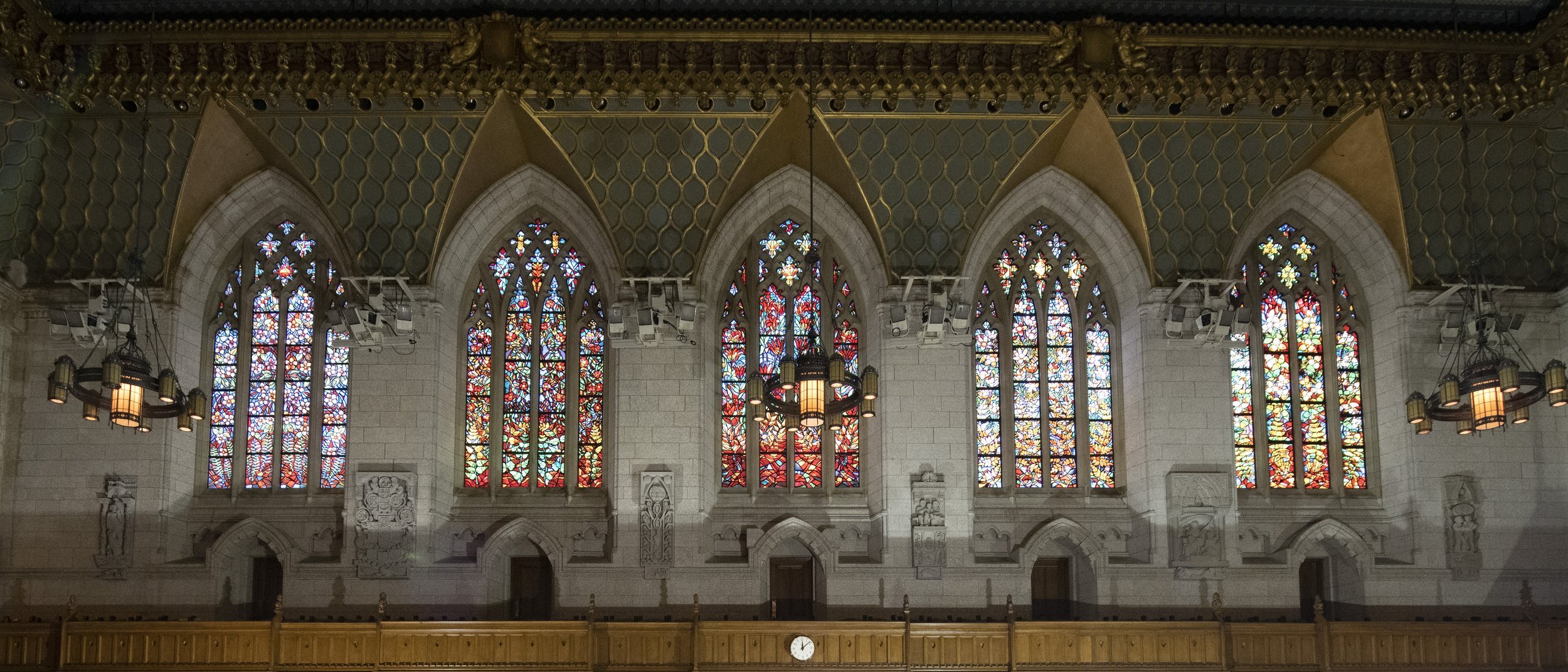 Interior of a church with five large stained glass windows, decorative stone walls with carved reliefs, hanging chandeliers, and a wooden balcony.