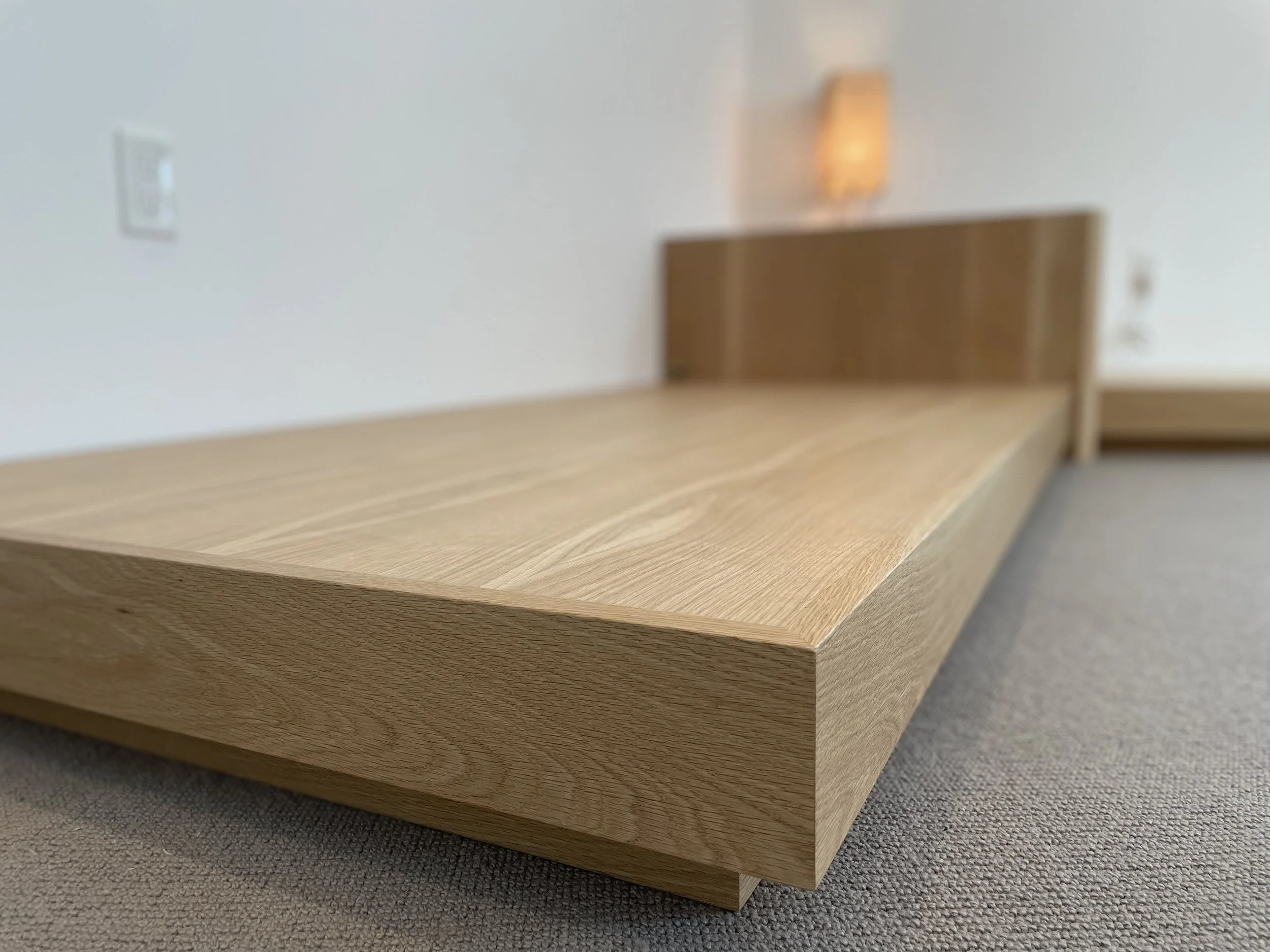 A low wooden platform bed with a headboard in a minimalistic room, with a textured gray carpet and white walls, featuring a wall-mounted light fixture.