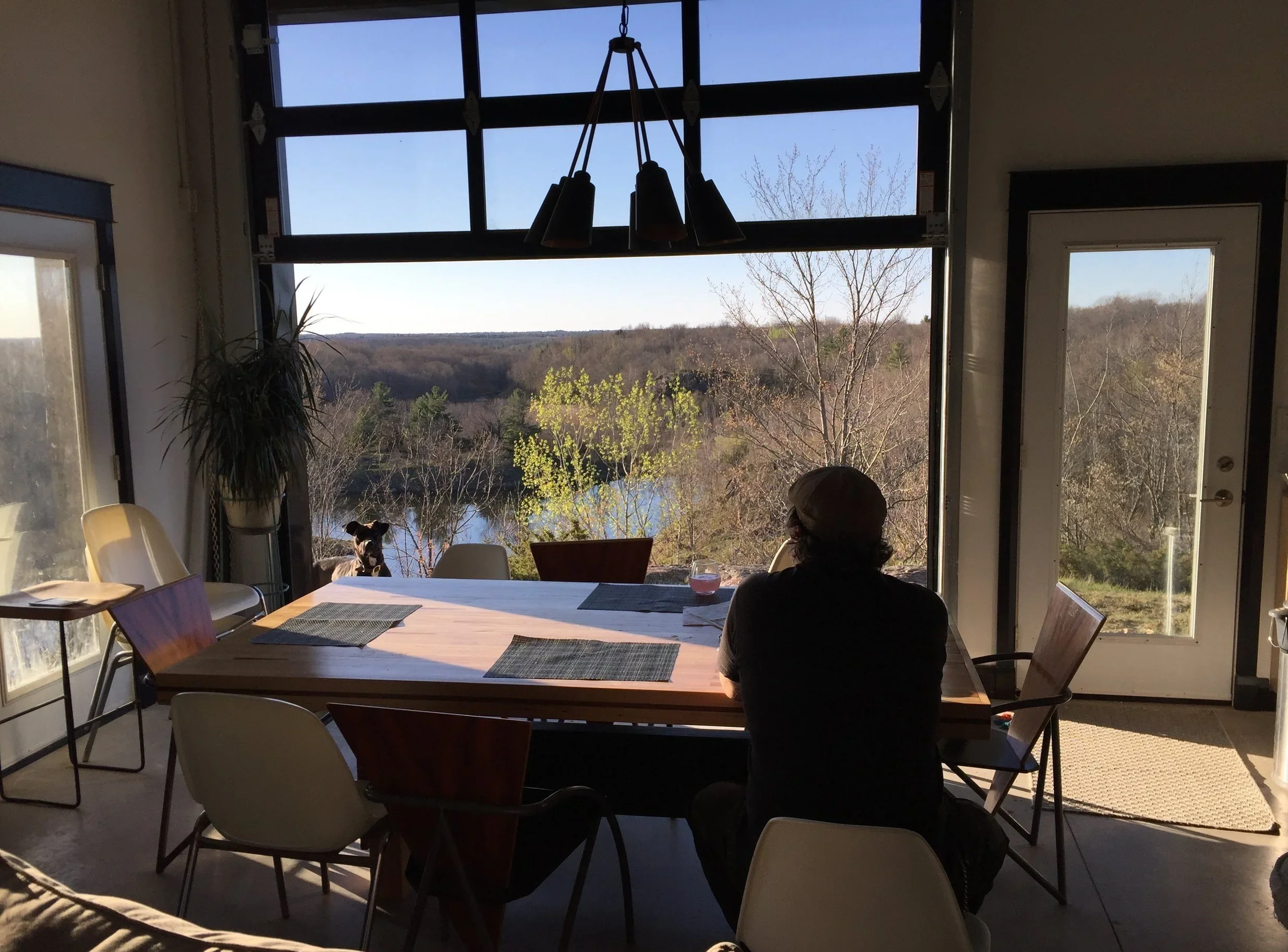 Interior of a house with a large window offering a view of trees and a river. A person with a hat is sitting at a dining table, looking outside. There is a small dog on the window sill. Some chairs, a hanging light fixture, and a plant are visible inside.