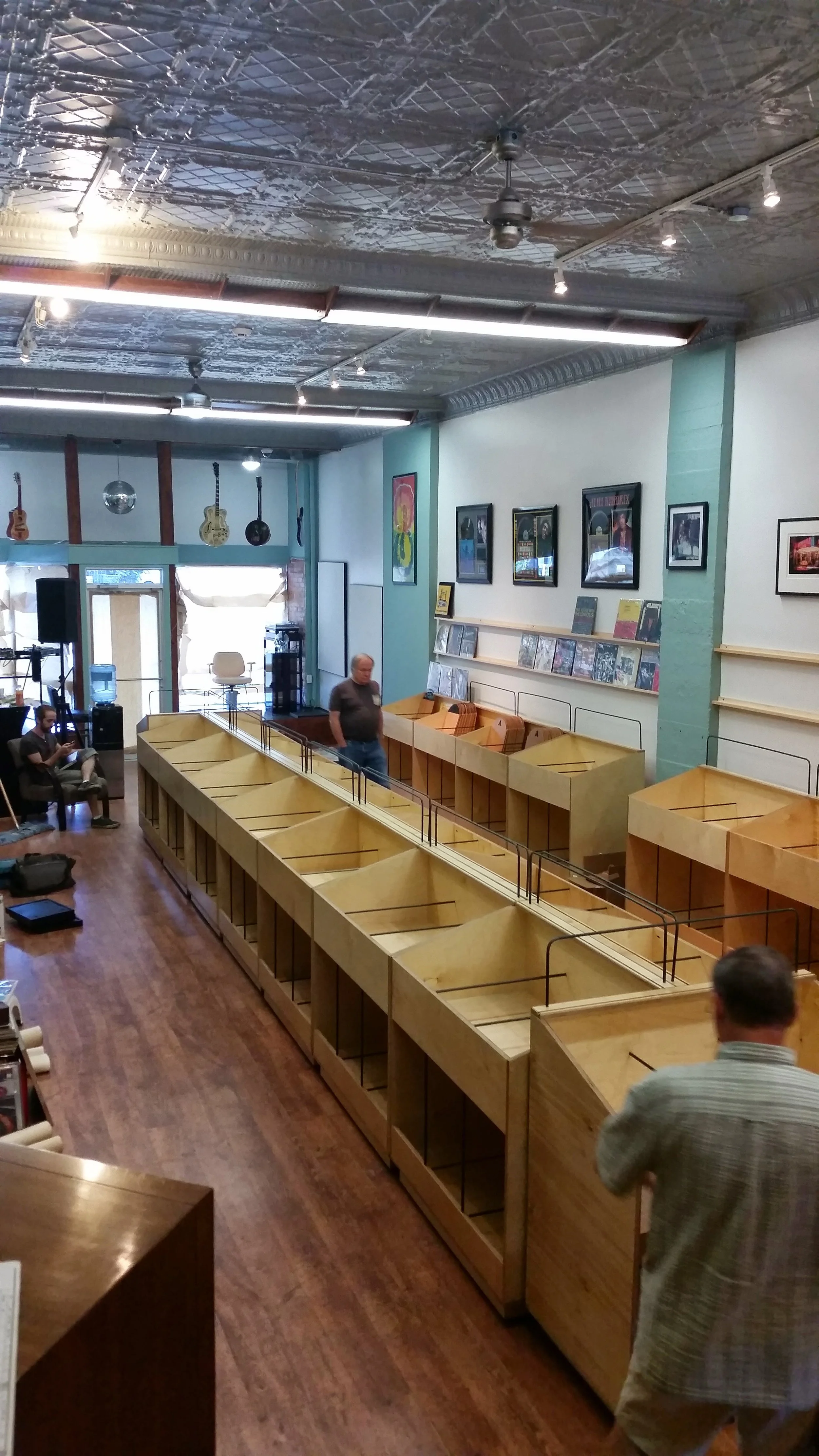 Interior of a record store with empty wooden bins, guitars hanging from the ceiling, framed pictures on the wall, and two people standing and sitting.