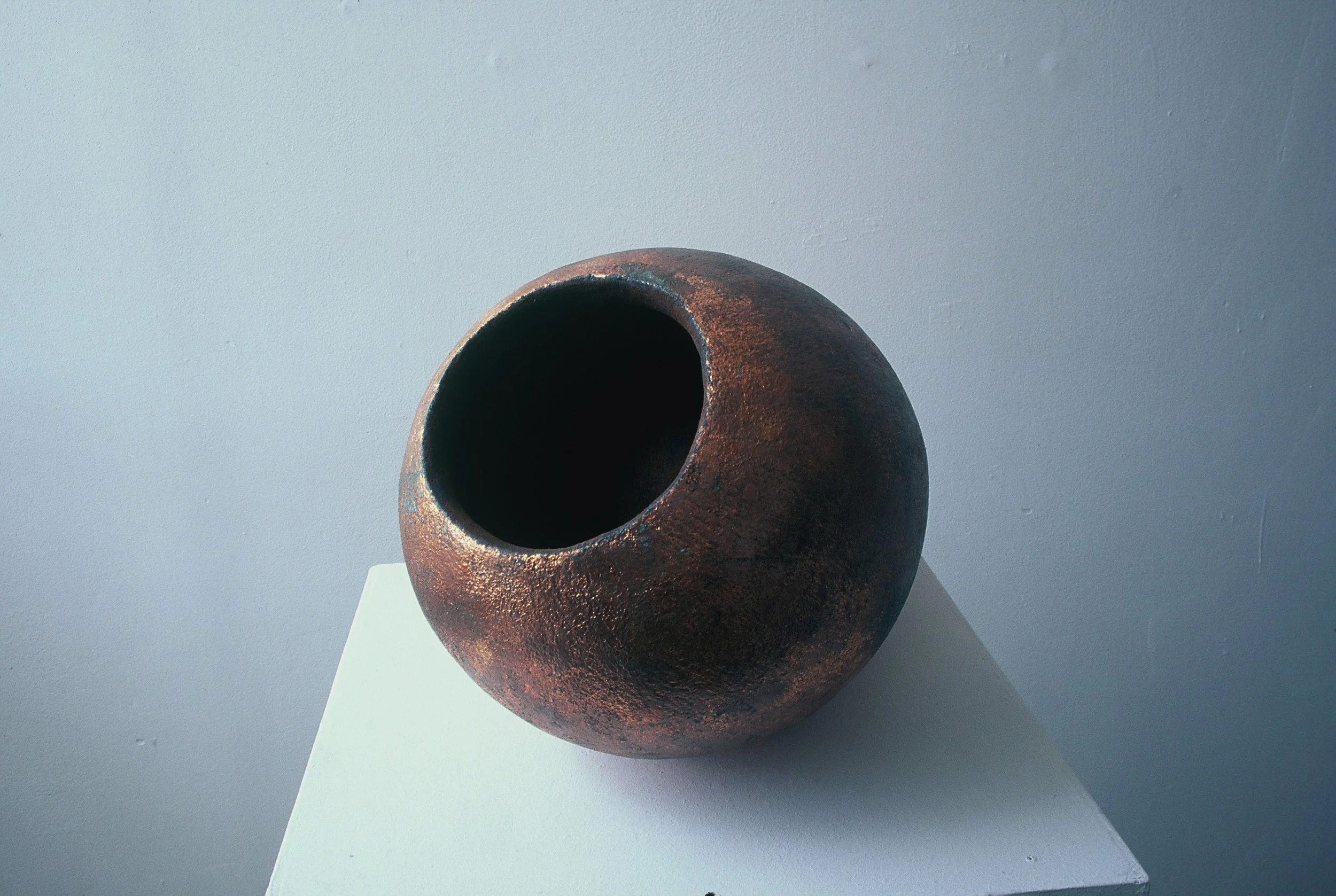 A round, rust-colored ceramic vase with a large, circular opening, placed on a white pedestal against a light blue wall.