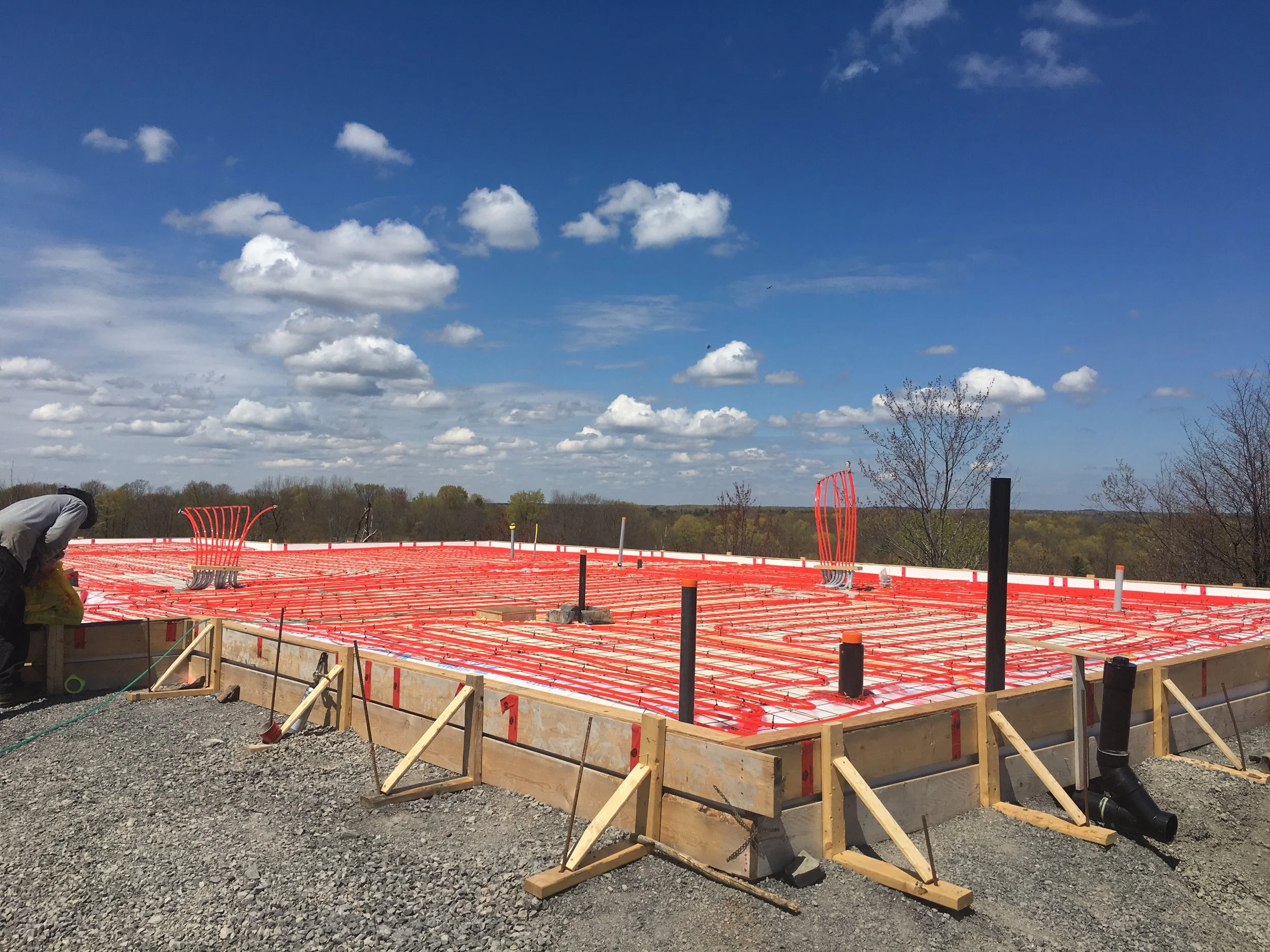 Construction site with a concrete foundation in progress, orange tubing and piping, and a worker on the left side, under a partly cloudy sky.