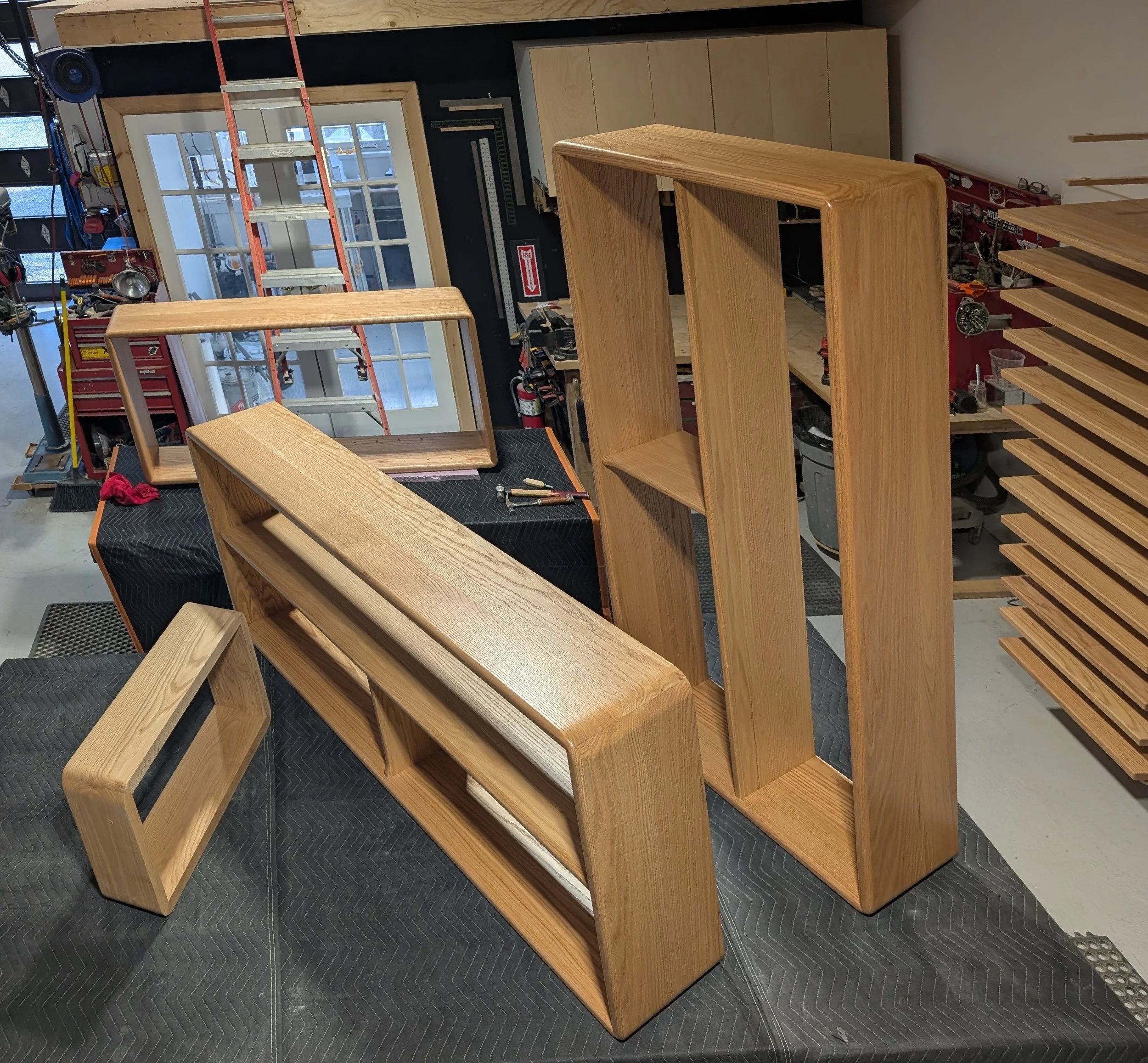 Unfinished wooden furniture pieces, including a rectangular open frame, a bench, a tall shelving unit, and a small rectangular frame, arranged on black mats in a woodworking workshop.