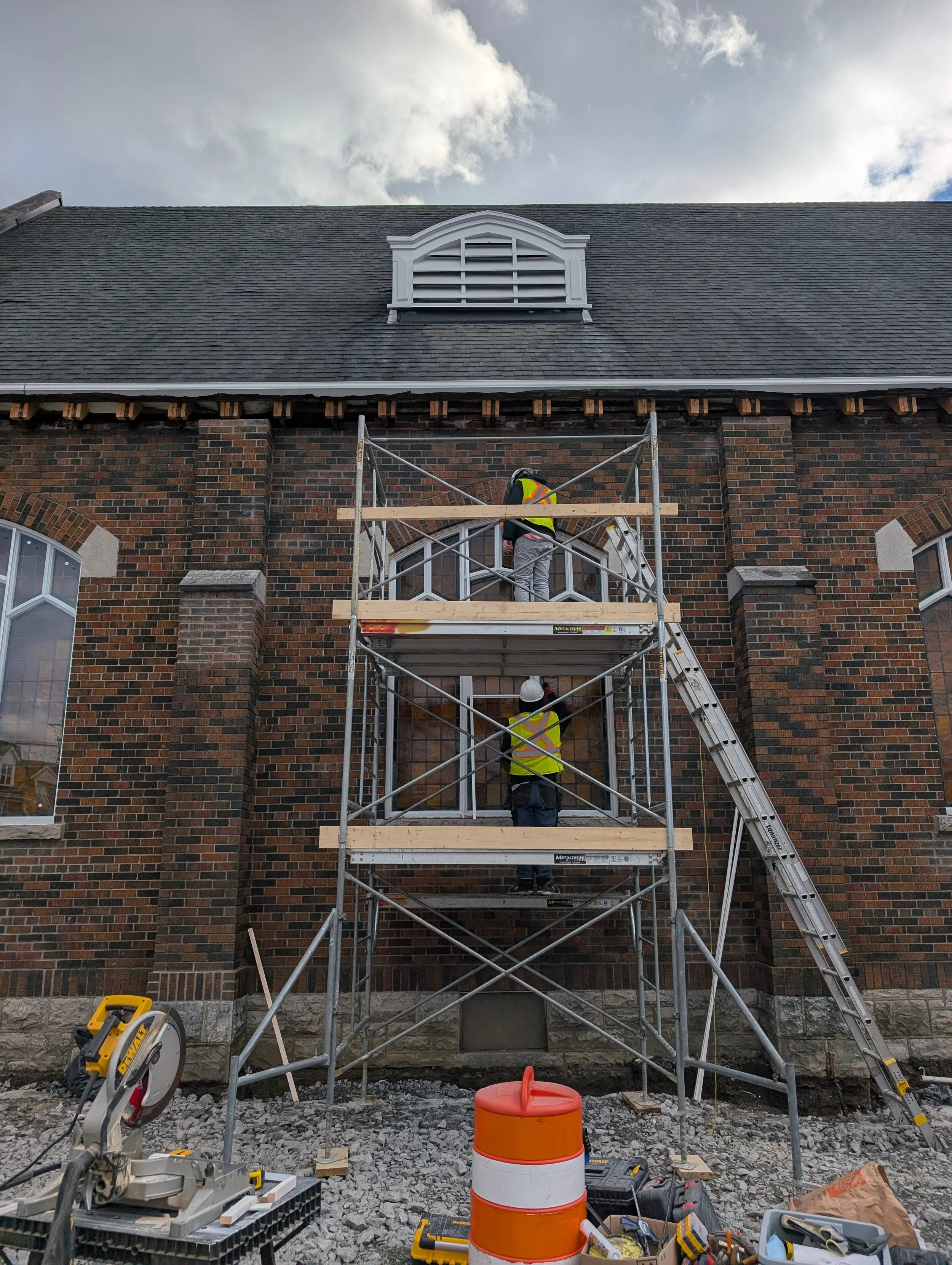 Construction workers in safety vests and helmets working on scaffolding outside a brick building with a window and attic vent, with construction tools and equipment on gravel ground.