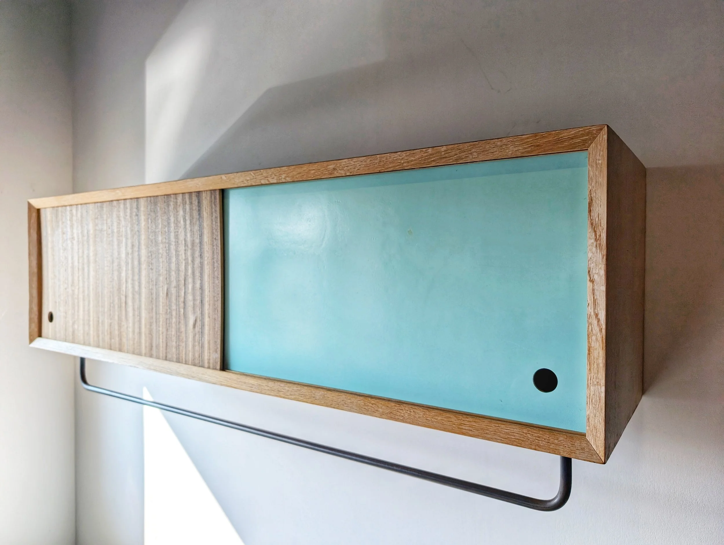 A rectangular wooden cabinet with sliding doors, one side woodgrain and the other painted light blue, mounted on a white wall with a black metal support underneath.