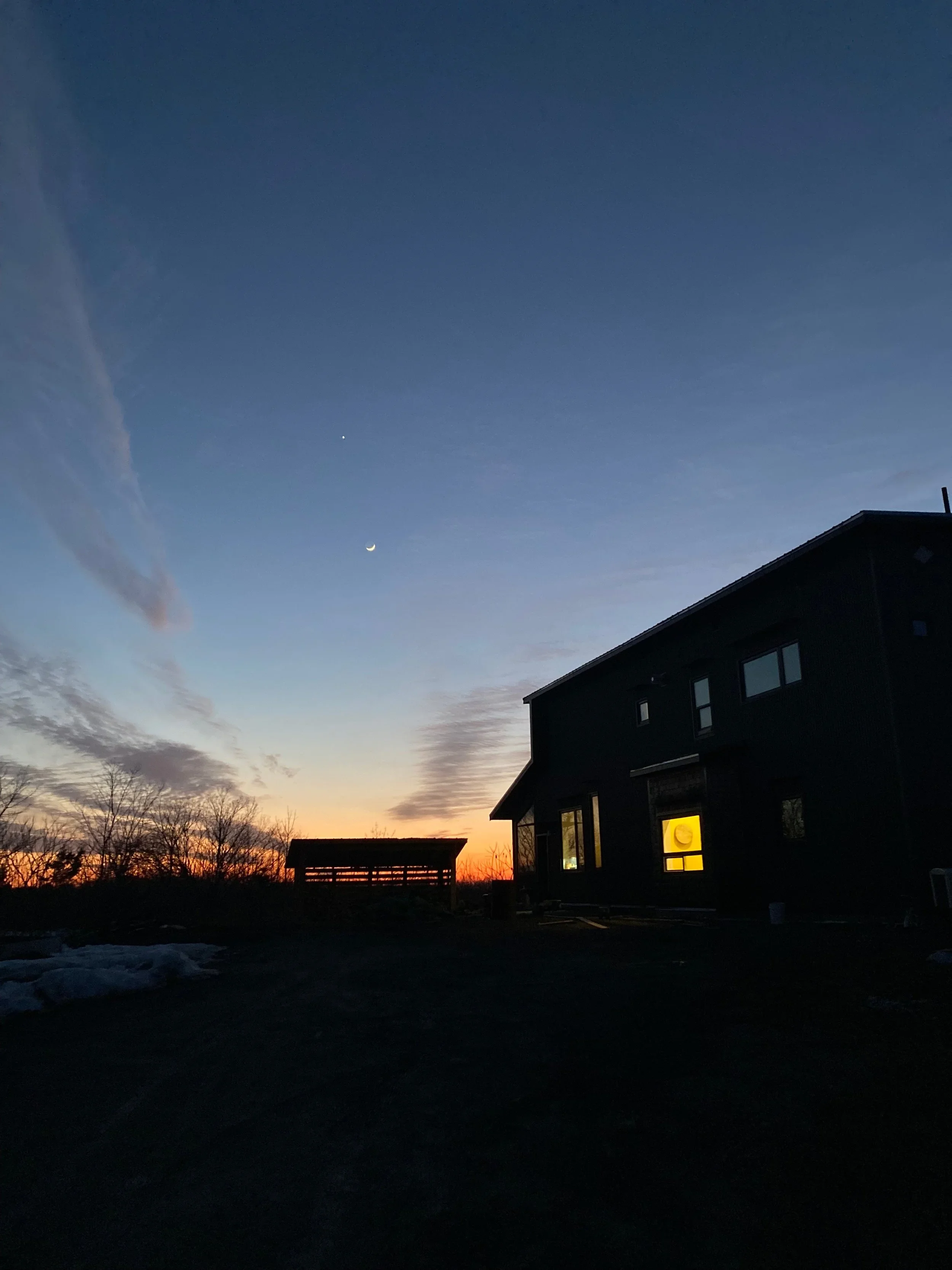 Dusk sky with visible moon and bright planet or star, dark house with lit window, and trees in the distance.