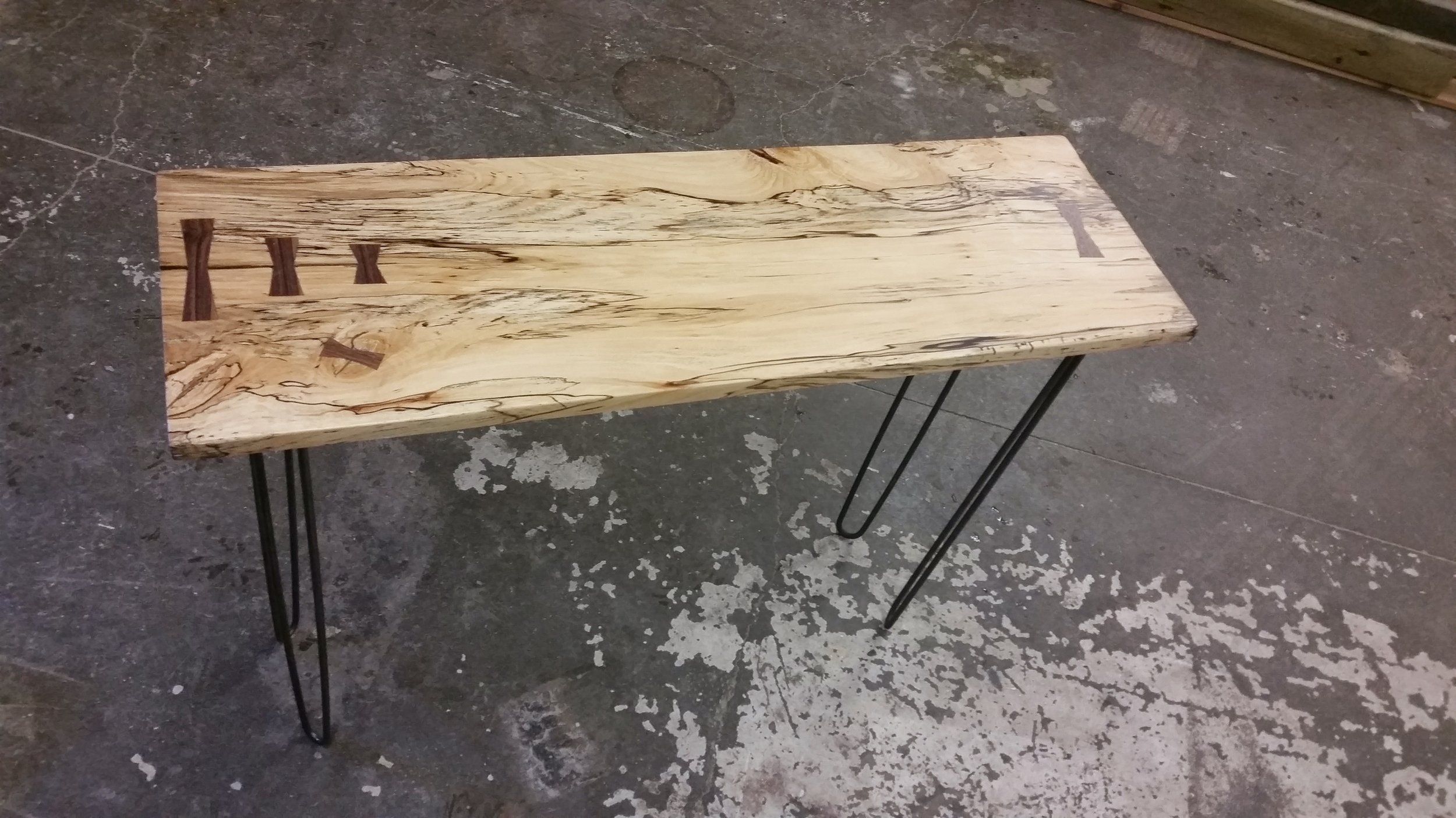 Wooden bench with a natural, live edge finish and hairpin metal legs, placed on a concrete floor.