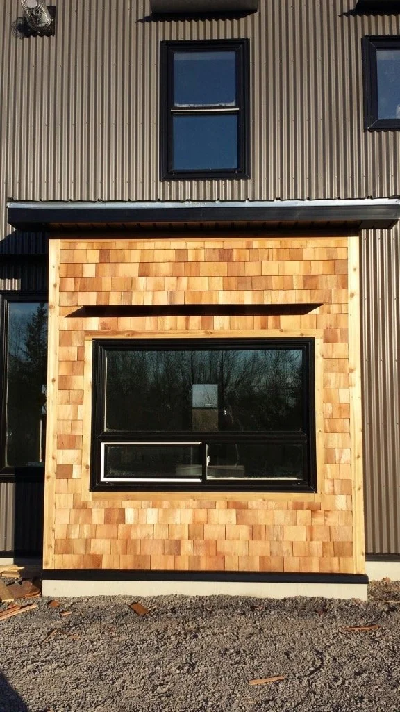 Close-up of a building exterior showing a black-framed window, wood shingles, and a metal siding wall.