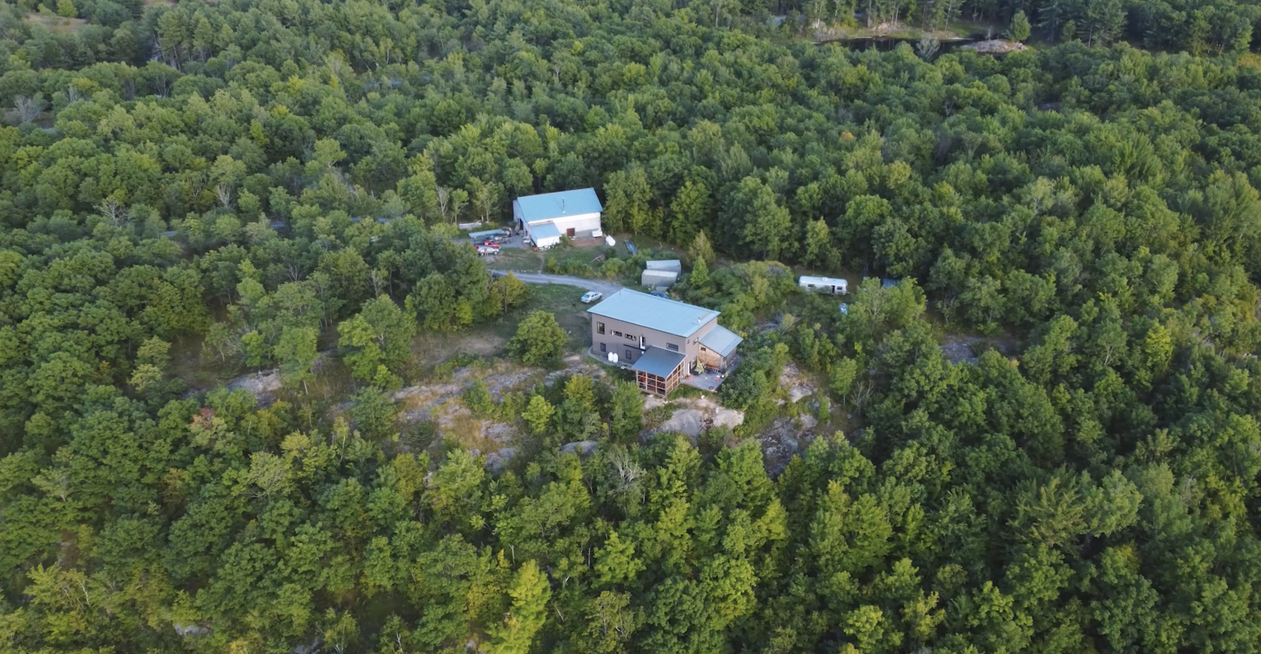 Aerial view of two houses surrounded by dense green trees, with a gravel driveway, parked cars, and trailers in a wooded area.