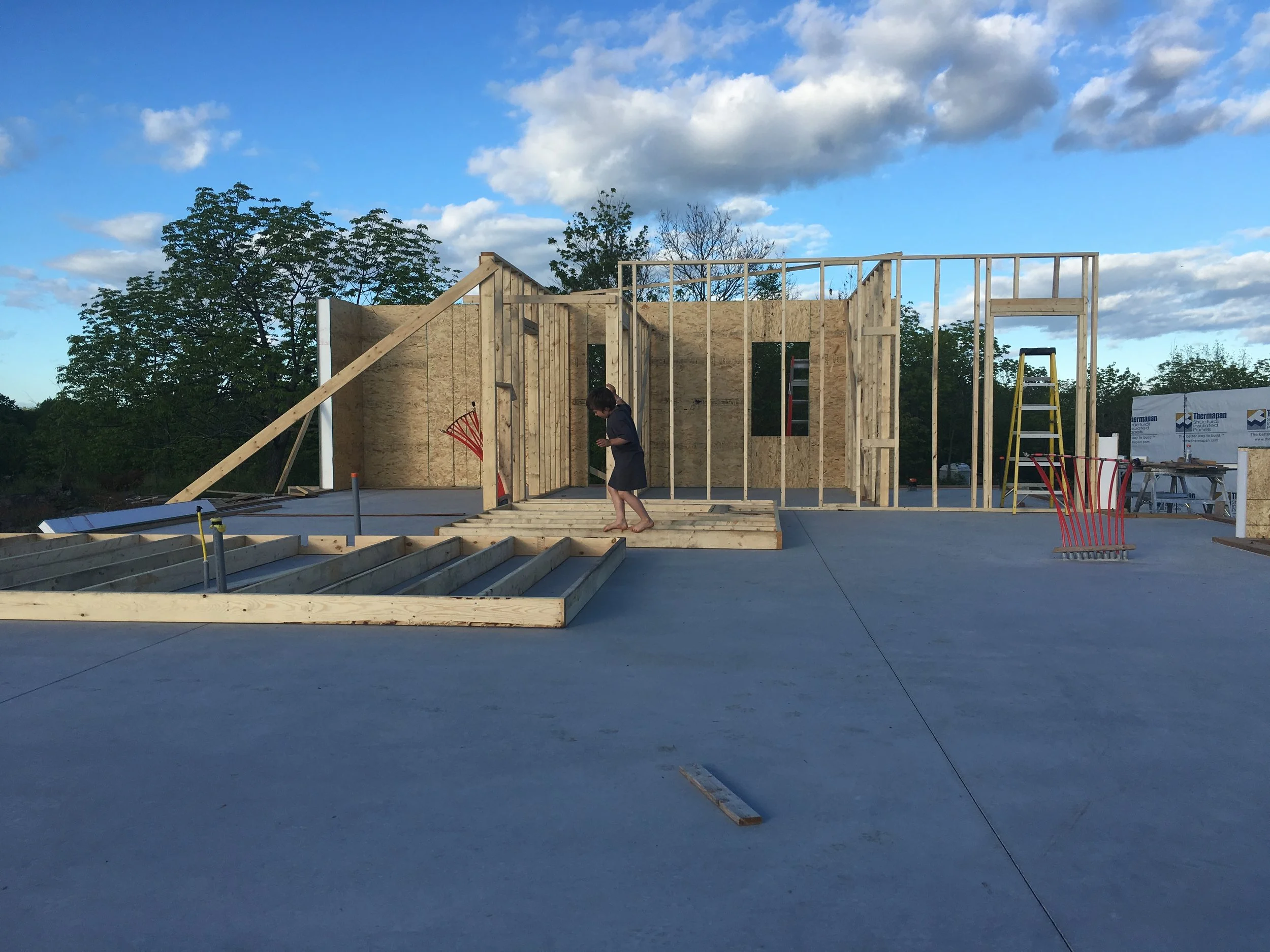 Wooden framing being constructed on a concrete foundation of a building, with a person walking on the foundation, on a clear day with clouds and trees in the background.