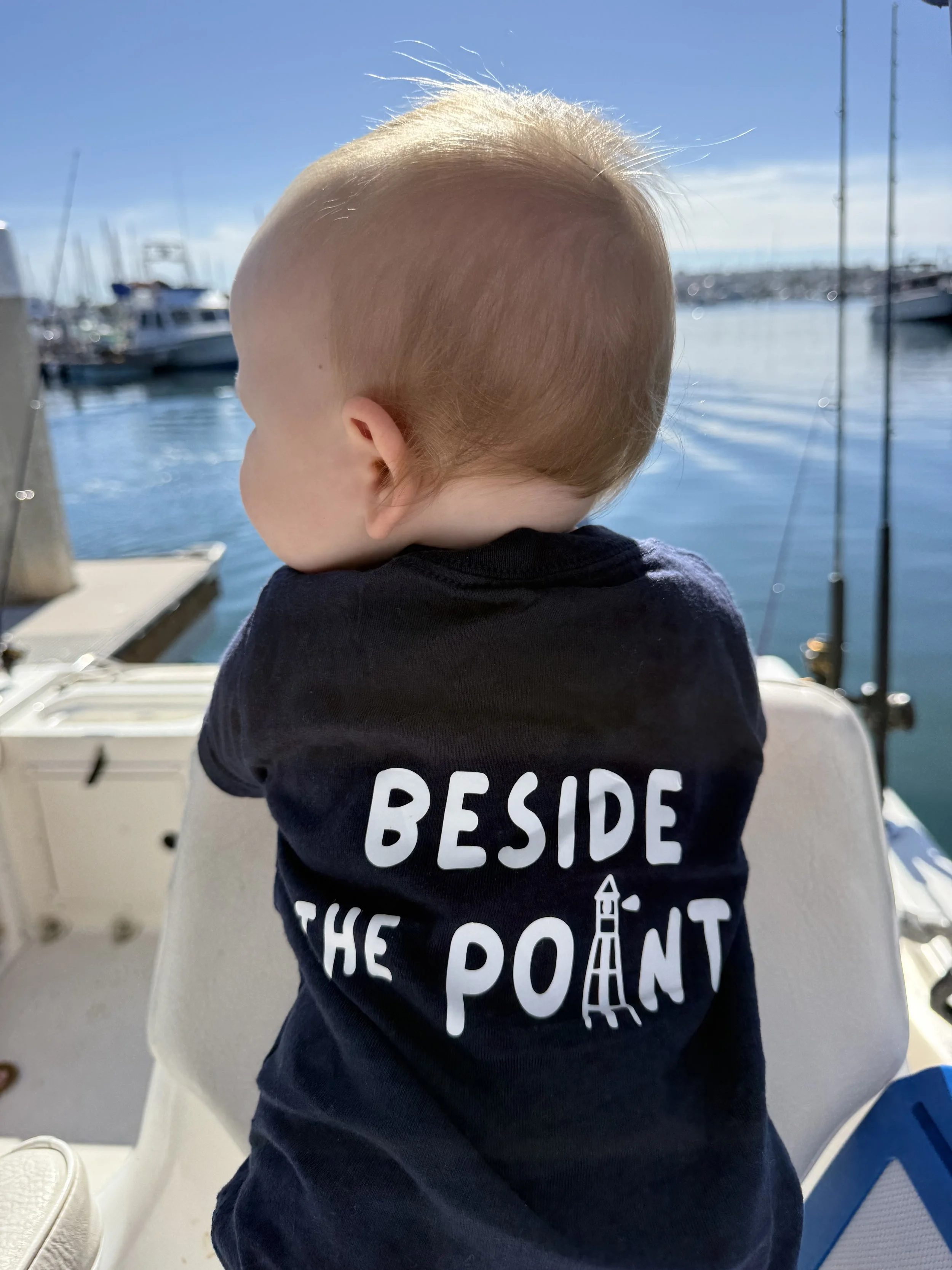 A young child sitting on a boat at a marina, wearing a navy T-shirt with the words 'BESIDE THE POINT' and a lighthouse graphic on the back, looking out at the water and boats. Beside the Point apparel for kids. Boat babies and young anglers love it!